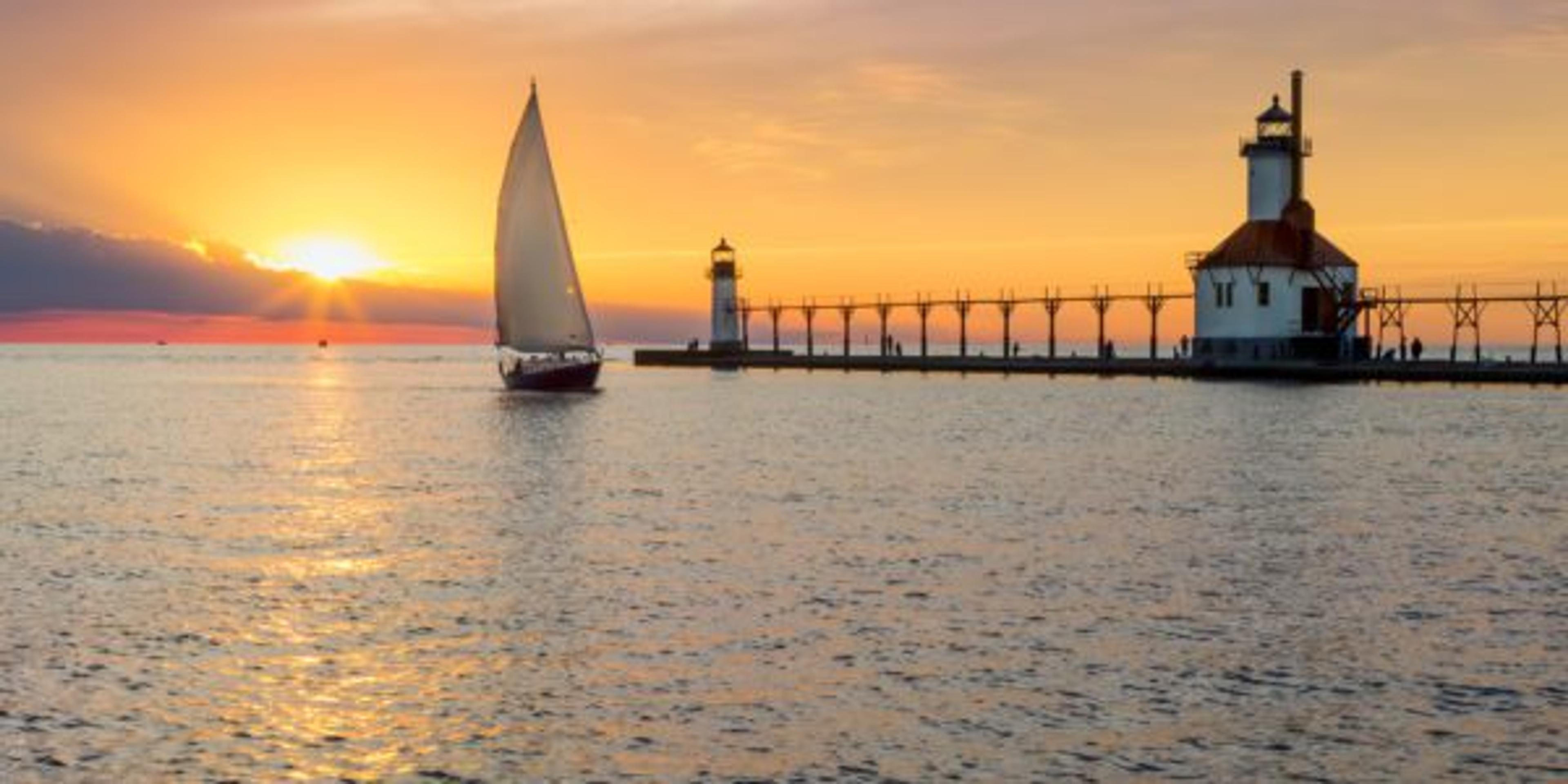 A sailboat rounds the breakwater near sunset on the longest day of the year by the Lighthouses at St. Joseph, Michigan with people fishing from and walking on the pier.