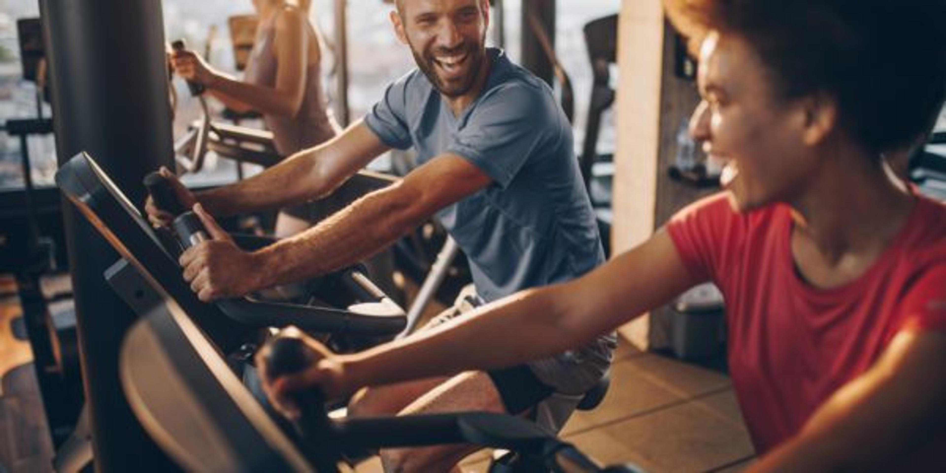 Man cheerfully rides a stationary bike while conversing with the woman beside him.