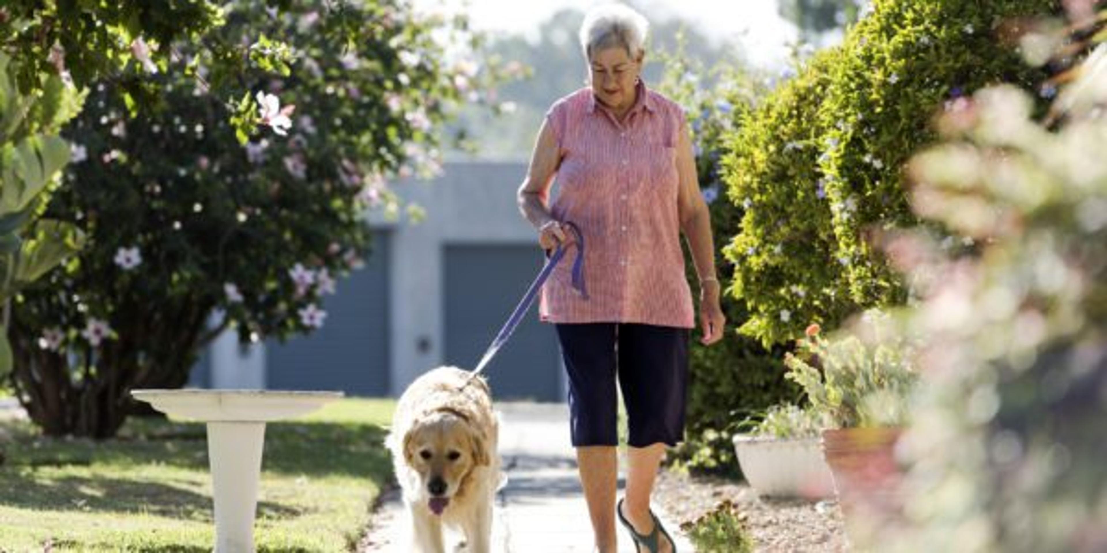 Photo of a smiling senior woman walking her dog outdoors along a foot path