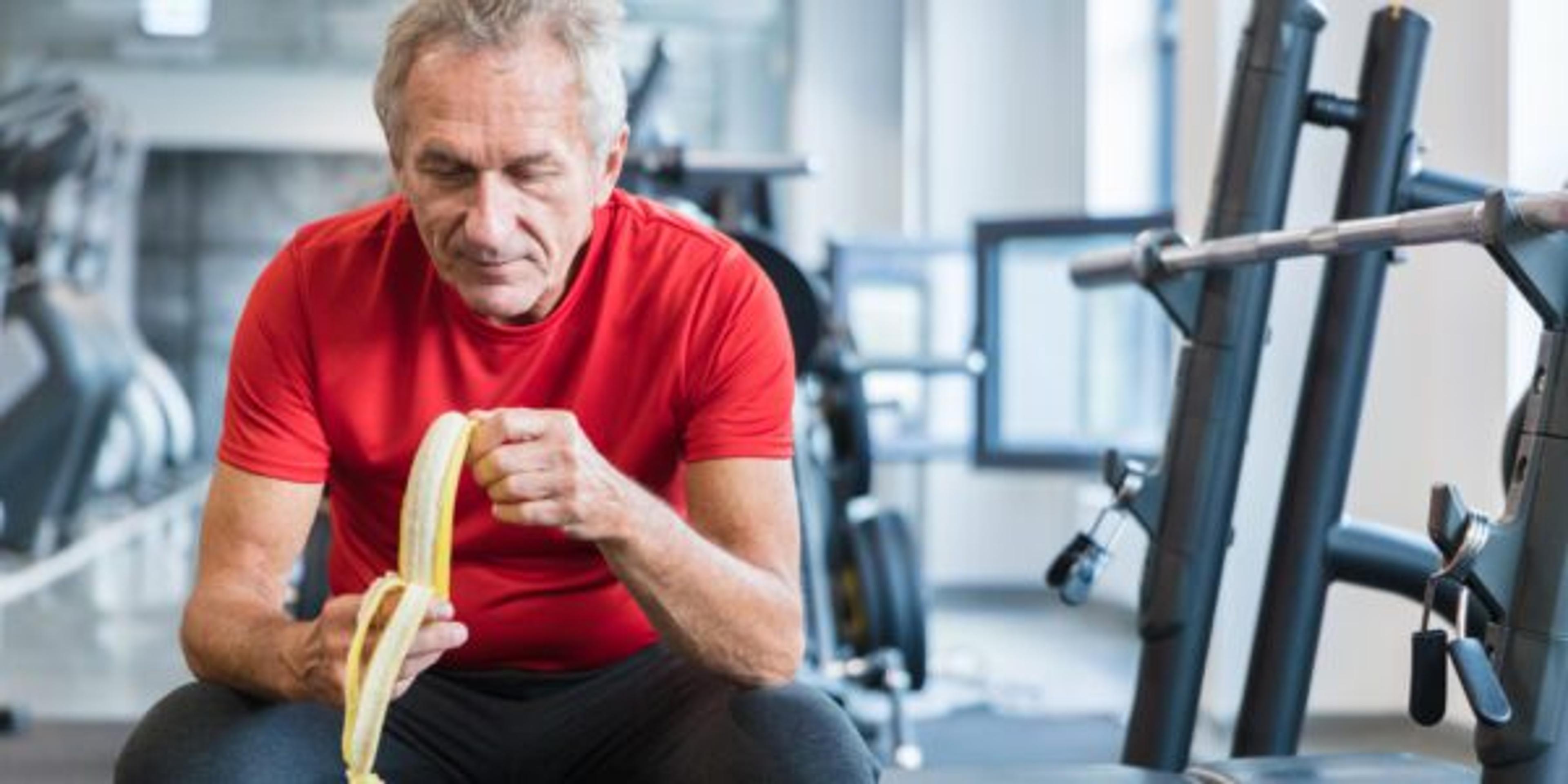 A man eats a banana while sitting on a weight bench at the gym.