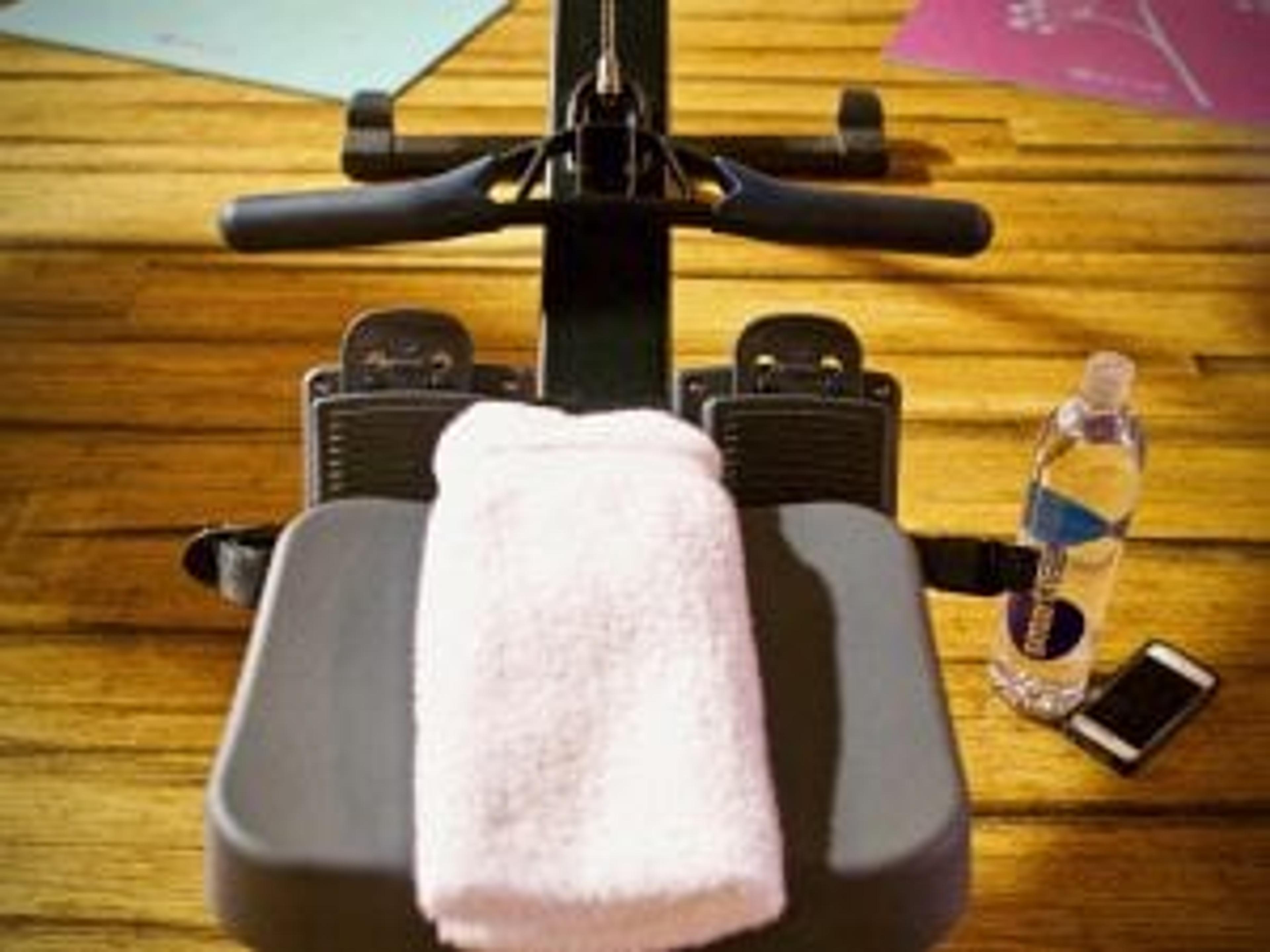 A towel rests on the seat of a rowing machine with a water bottle and phone placed on the floor to the right of the machine.