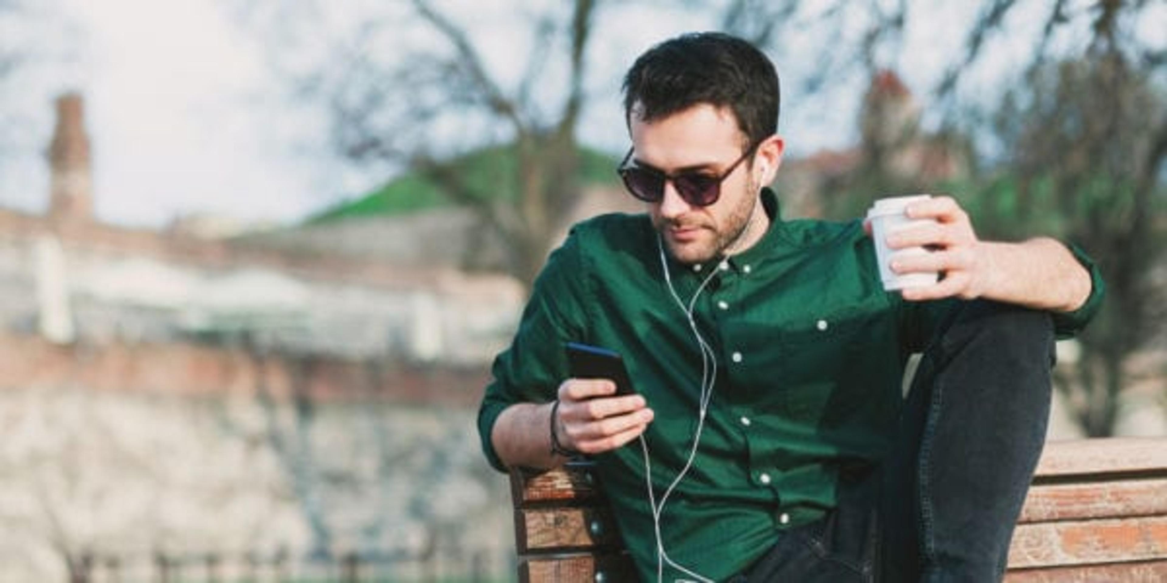 Young man drinking coffee listening to music on his smartphone through earbuds.