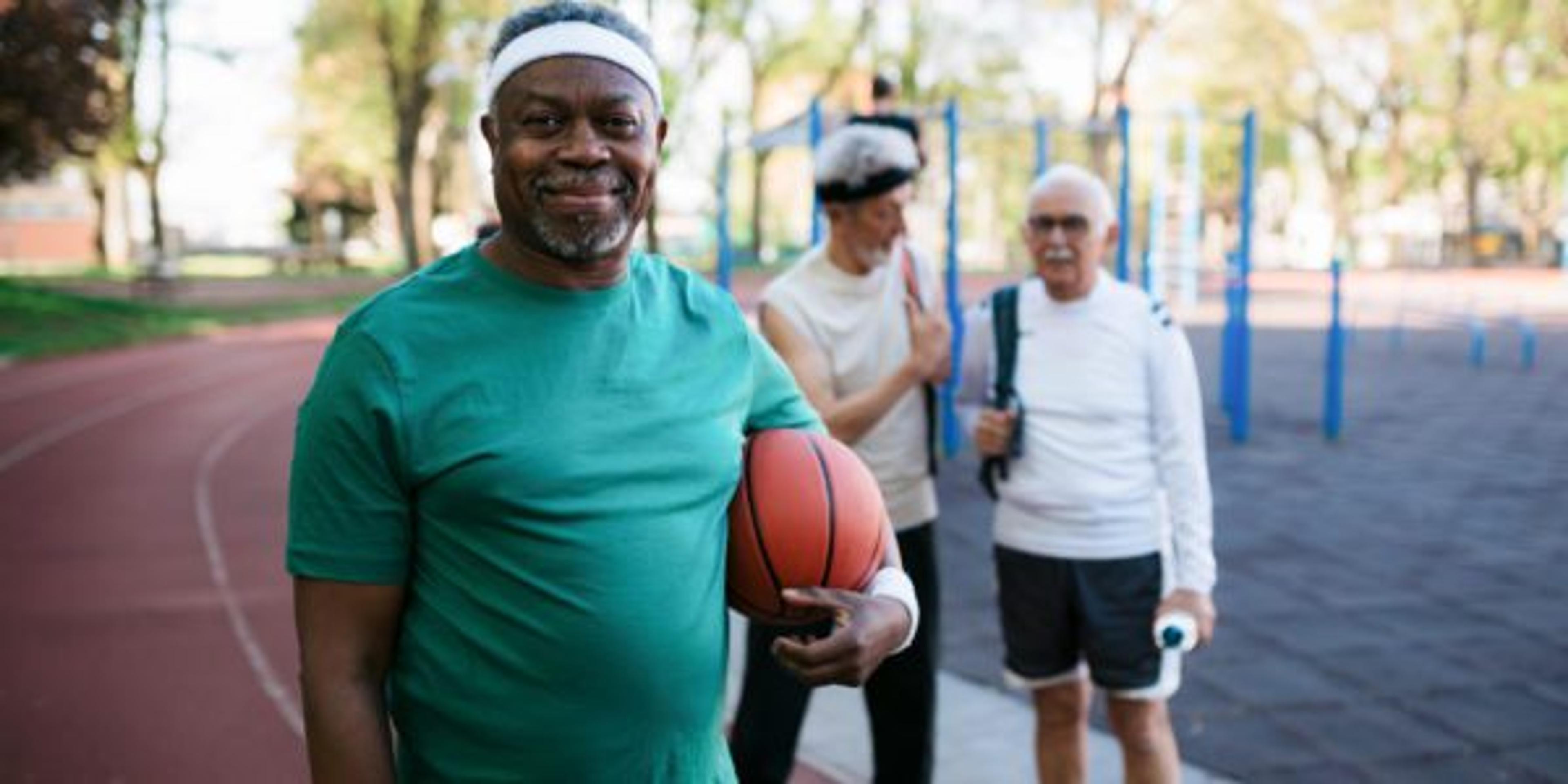 A Black man smiles at the camera holding a basketball as his friends look on from the park behind him.