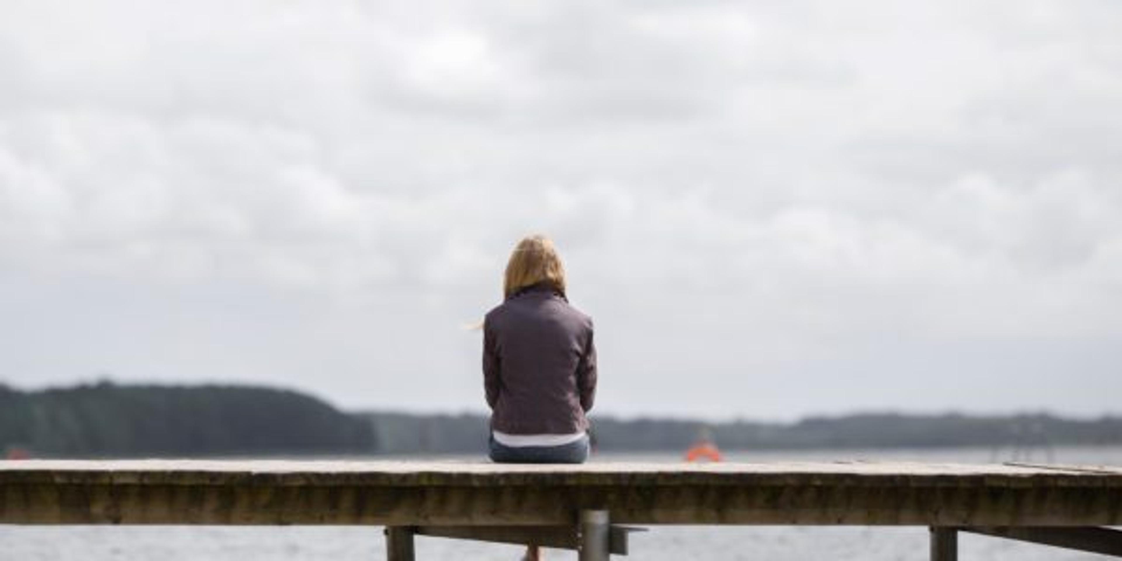 One young woman sitting alone on edge of footbridge and staring at lake and cloudy sky in summer day. Thinking about life. Spending time alone in nature. Peaceful atmosphere. Back view.
