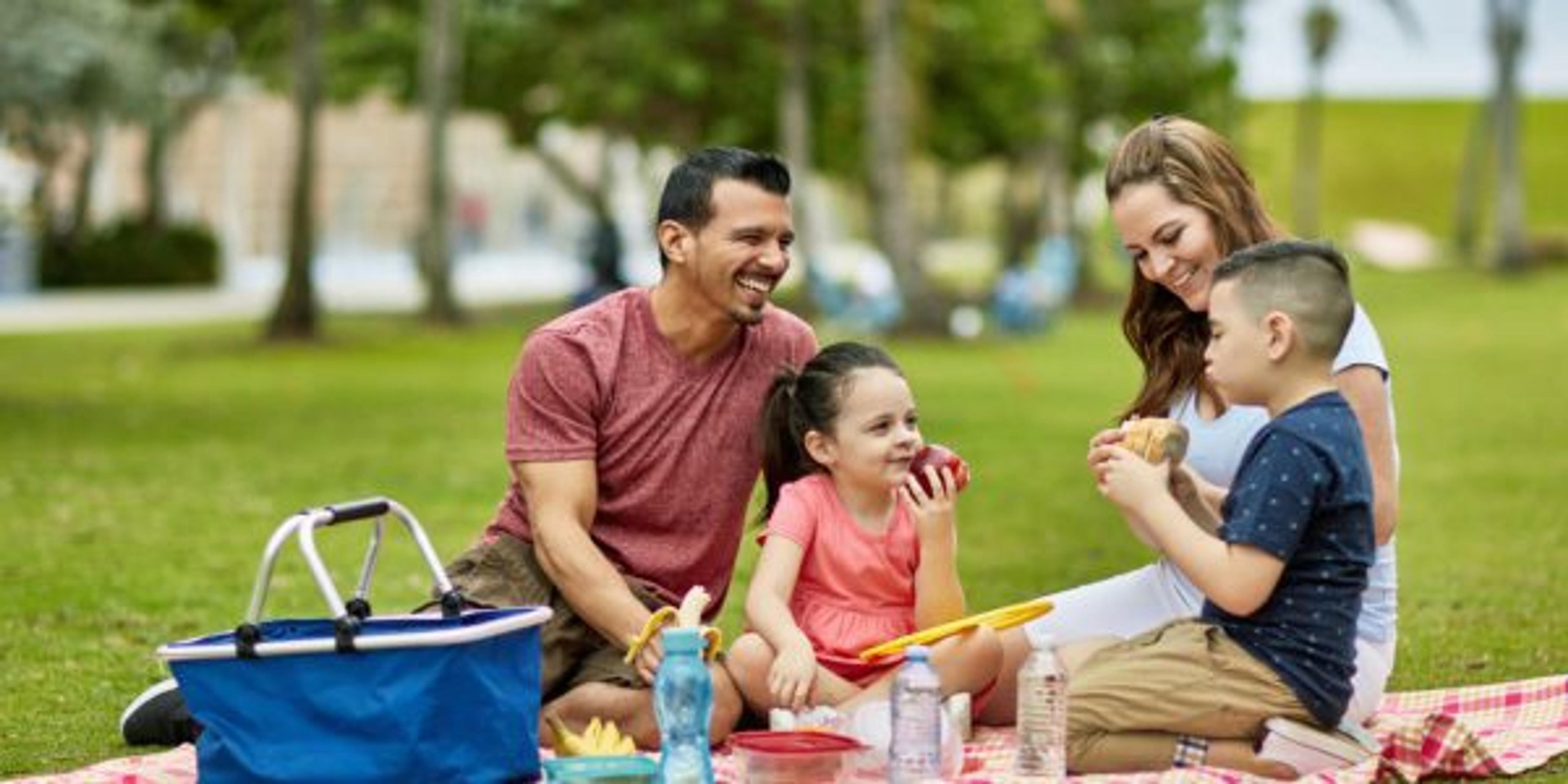 Hispanic American parents in late 30s enjoying picnic lunch and family time with their young children at Miami public park.