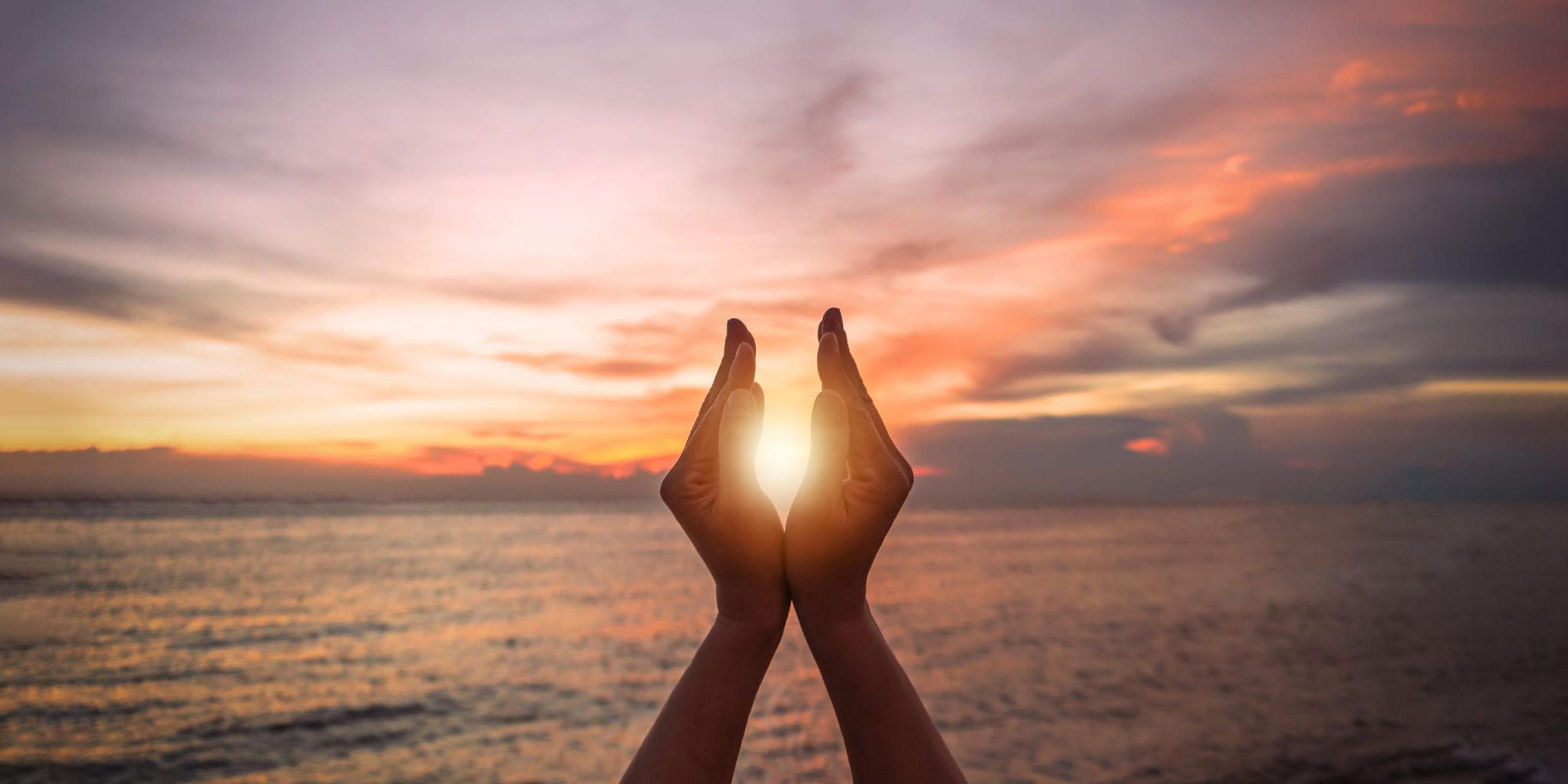 June summer sun solstice concept with silhouette of happy young woman's hands relaxing, meditating and holding sunset against warm golden hour sky on the beach with natural ocean or sea background