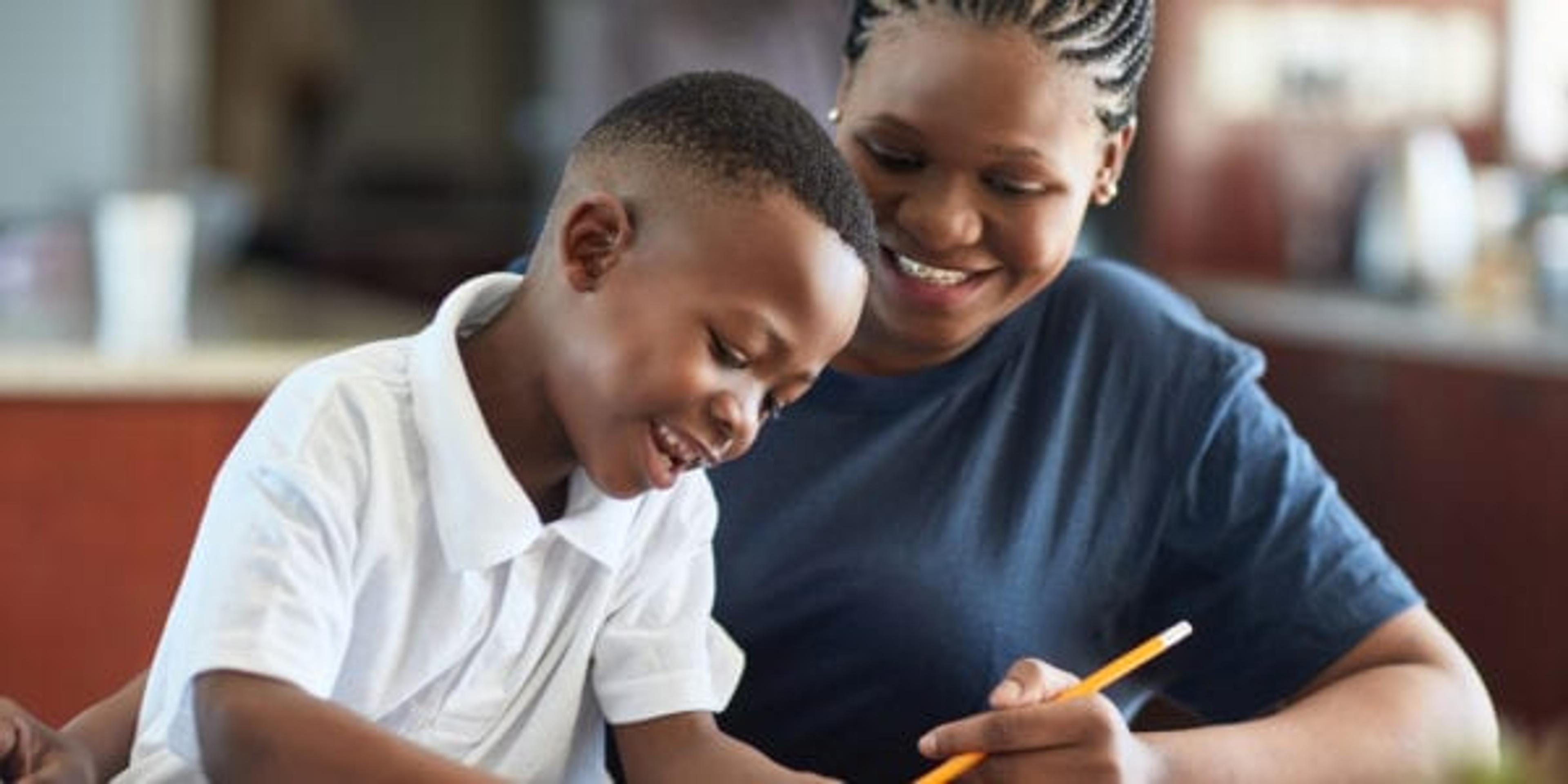 A Black mother and son sit at a desk doing schoolwork and smiling.
