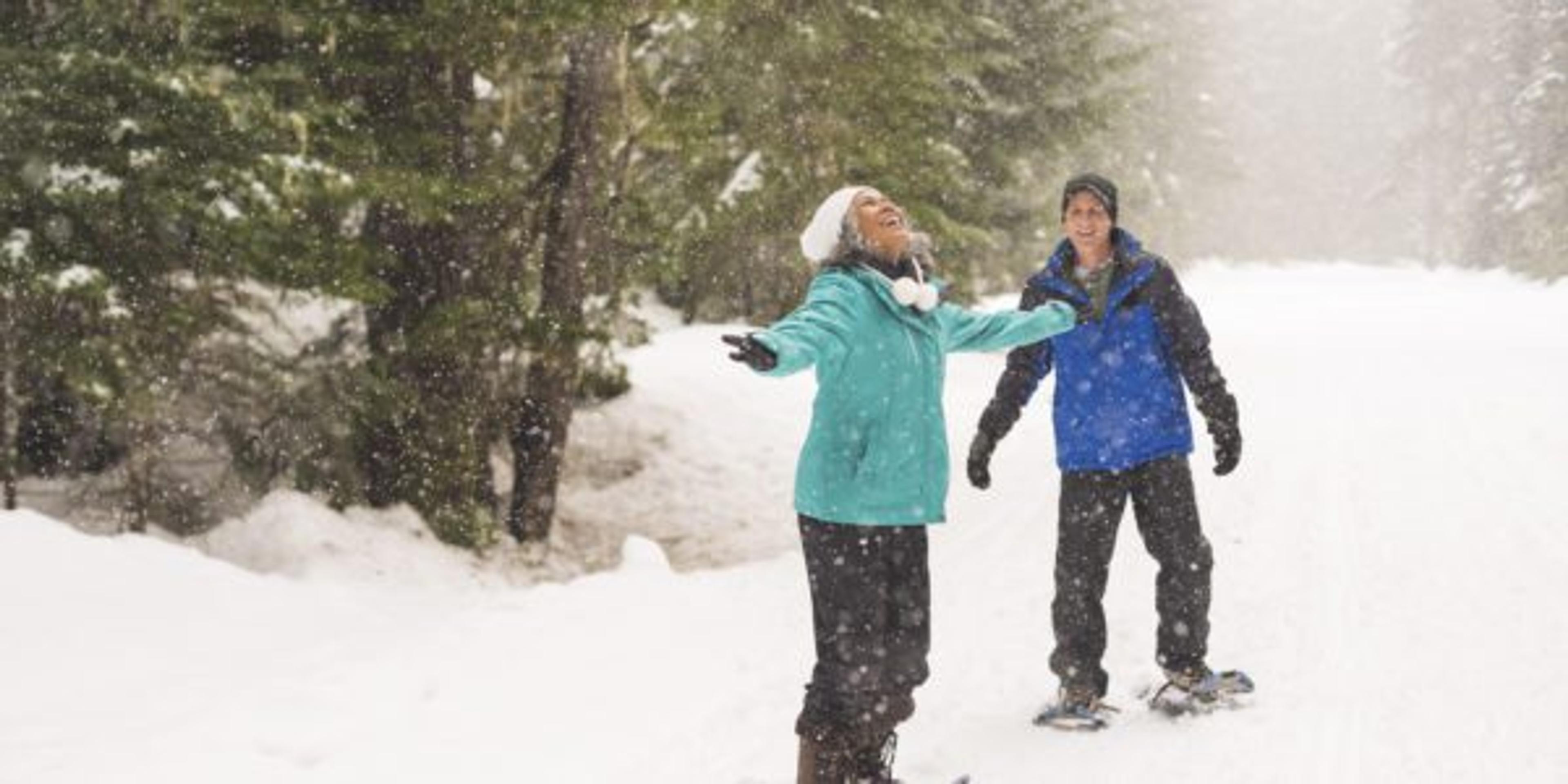 A mixed race senior couple joyfully embrace a snow shower while out for a walk in the forest. They are wearing snowshoes and warm clothing. The woman has her arms raised and is looking up at the falling snowflakes. The woman's husband is in the background walking towards her. He is smiling and watching her joyous display.