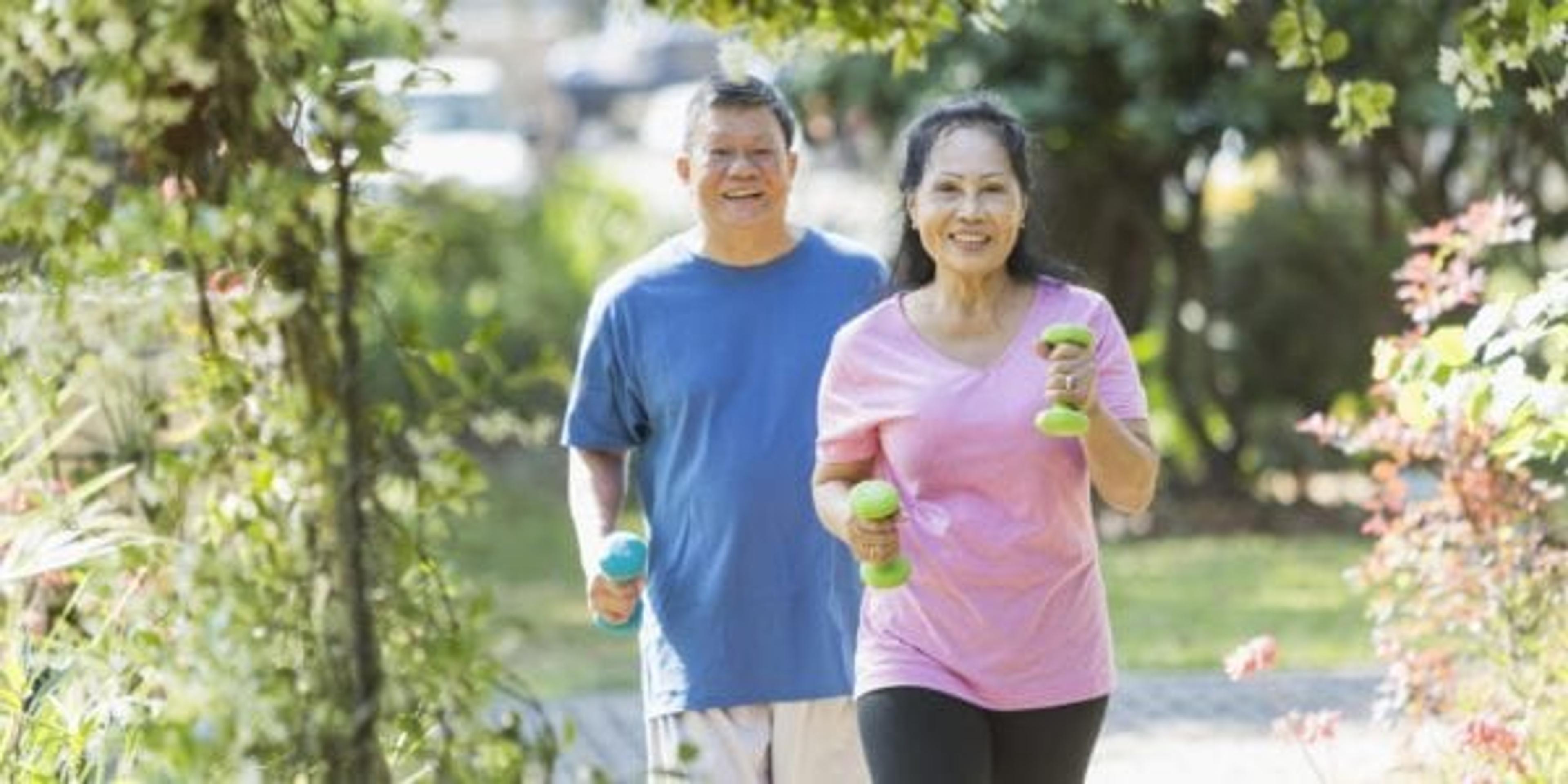 A senior Asian couple in the park on a sunny day, exercising together. They are powerwalking or jogging with dumbbells in their hands.