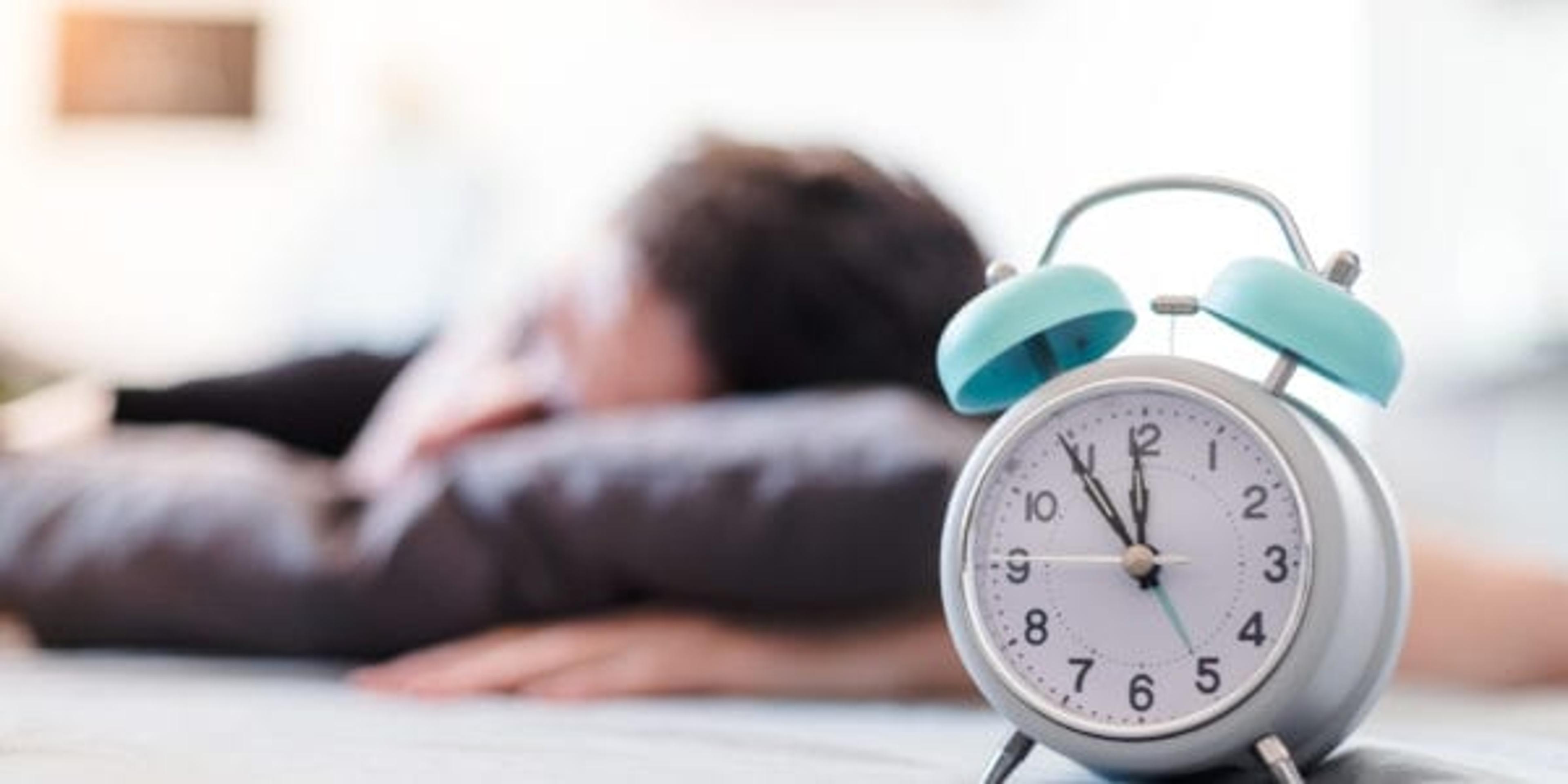 White alarm clock in the morning. Young man sleeps in the background.