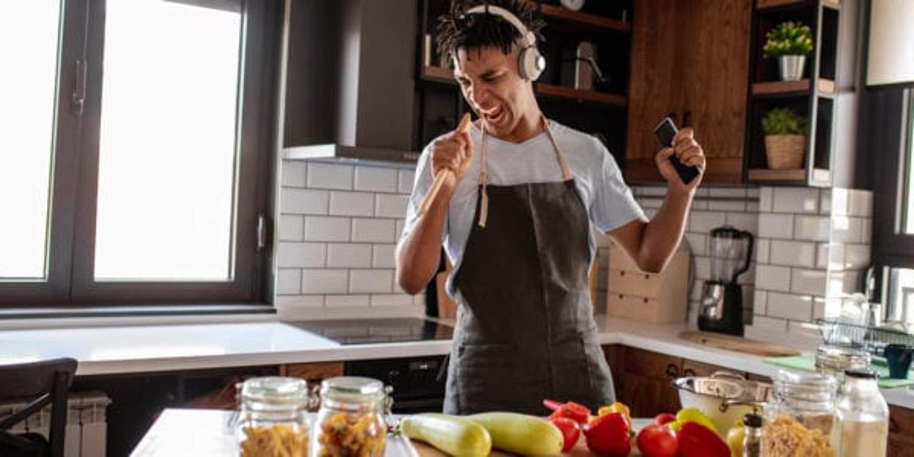 A young man is in the kitchen, he has wireless headphones on, he is dancing and singing