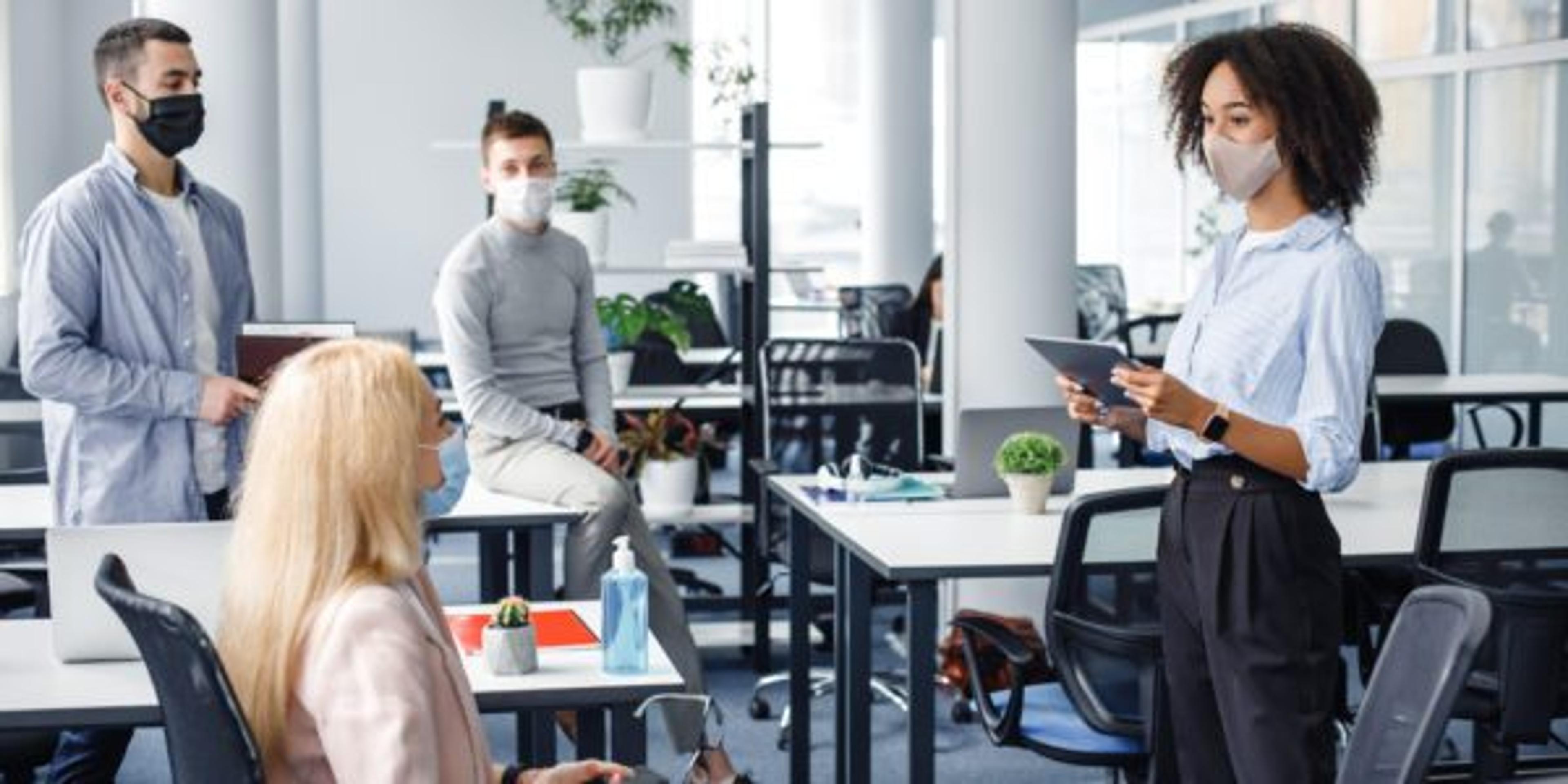 Corporate meeting and group work in company. African american woman manager in protective mask holding tablet, talking to workers keeping social distance