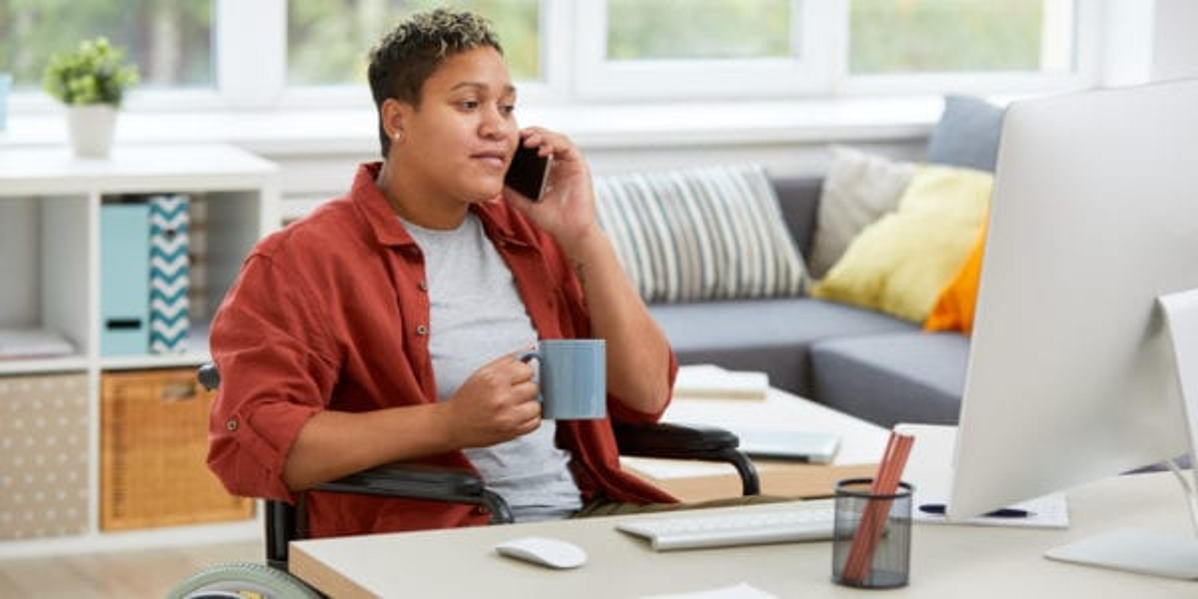 African disabled woman sitting in wheelchair at the table and looking at computer monitor while talking on mobile phone and drinking coffee at home