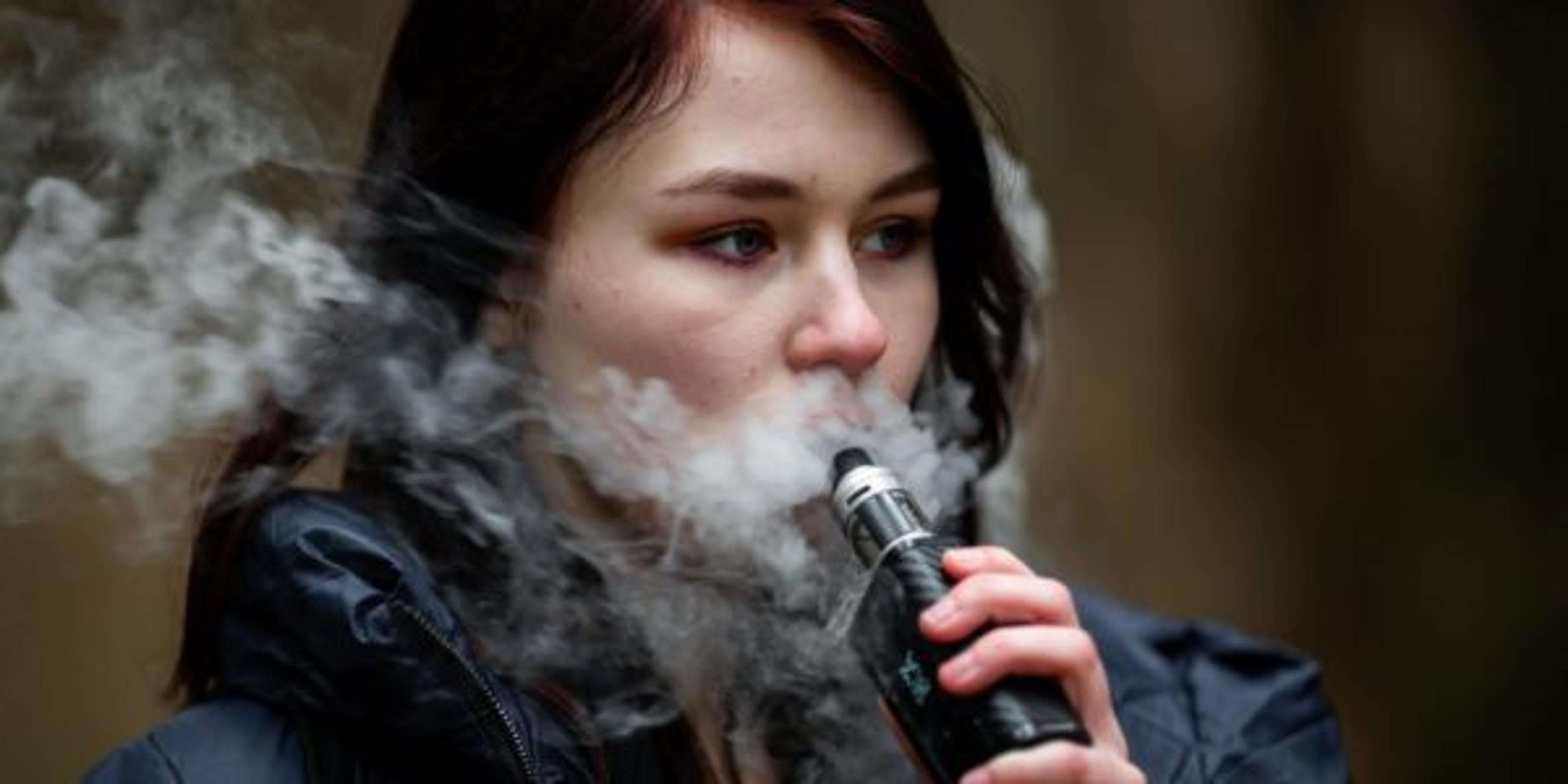 Vape teenager. Young pretty caucasian brunette girl smoking an electronic cigarette on the street in the spring. Bad habit.