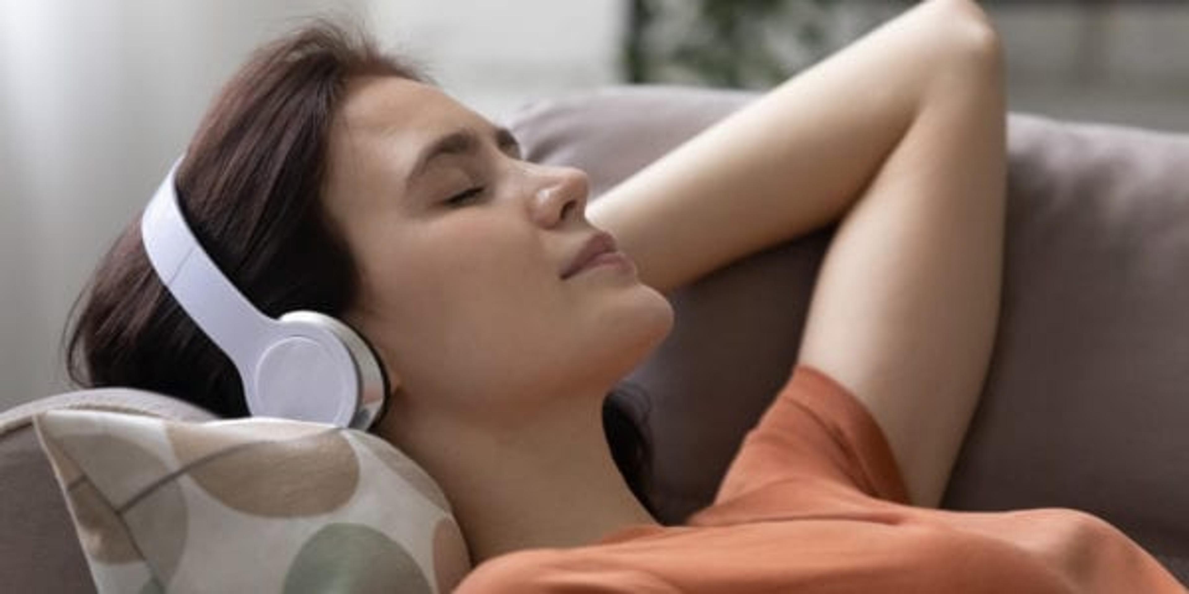 Head shot peaceful young woman lying on pillows on comfortable sofa, listening music or positive affirmations in modern headphones with closed eyes, relaxing alone at home, domestic hobby concept.