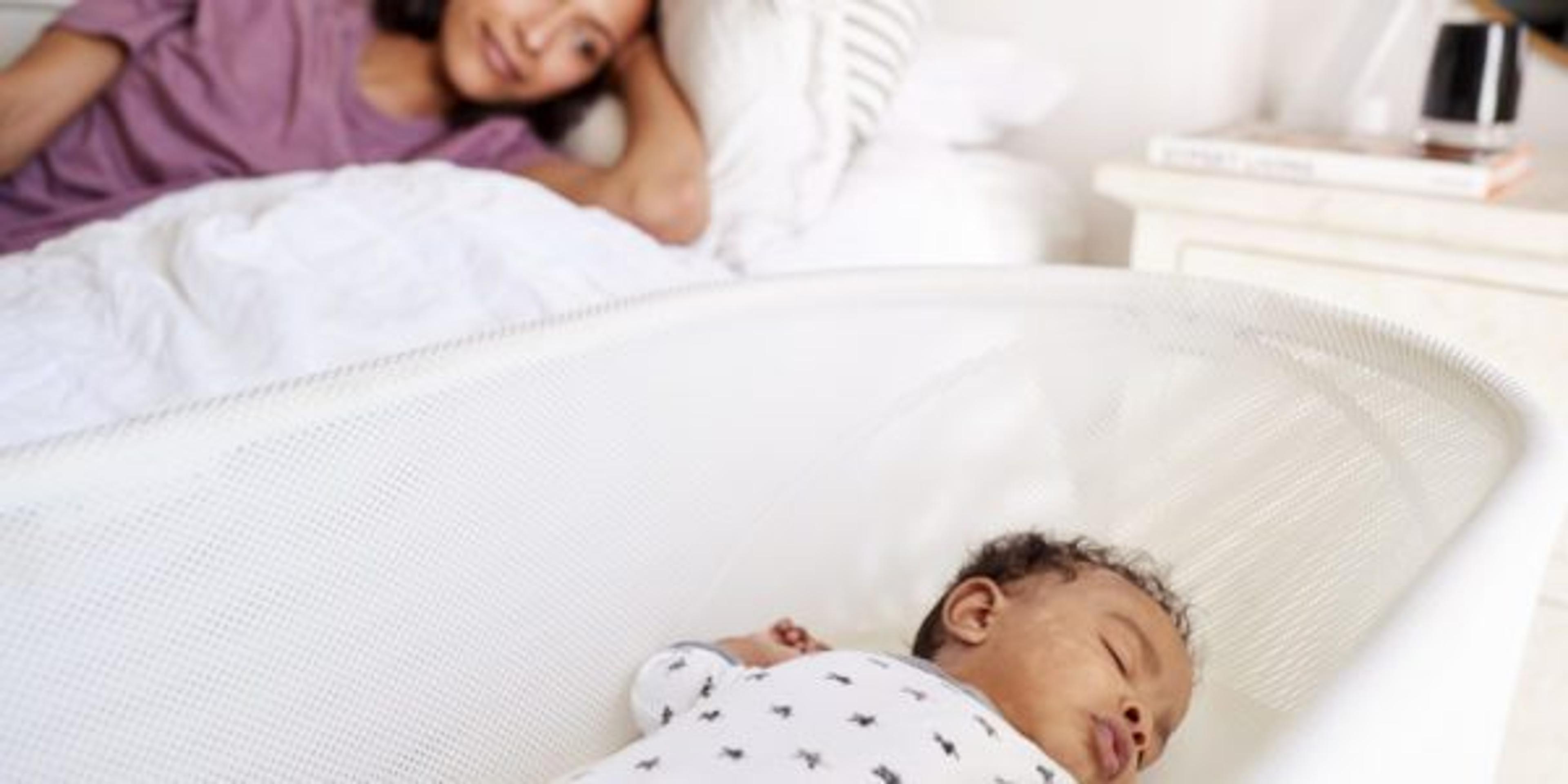 Young woman rests as her baby safely sleeps in her bassinet next to the bed