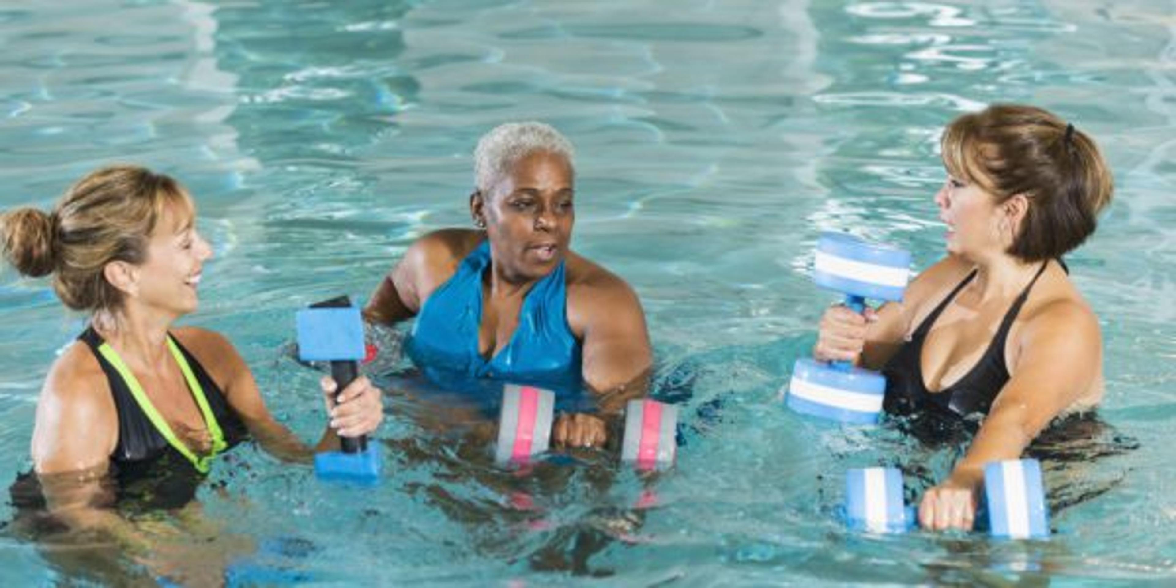 Multi-ethnic group of three middle aged and senior women (50s, 60s) doing water aerobics exercises with dumbbells in a swimming pool. Two of the women have serious expressions on their faces and the third is smiling.