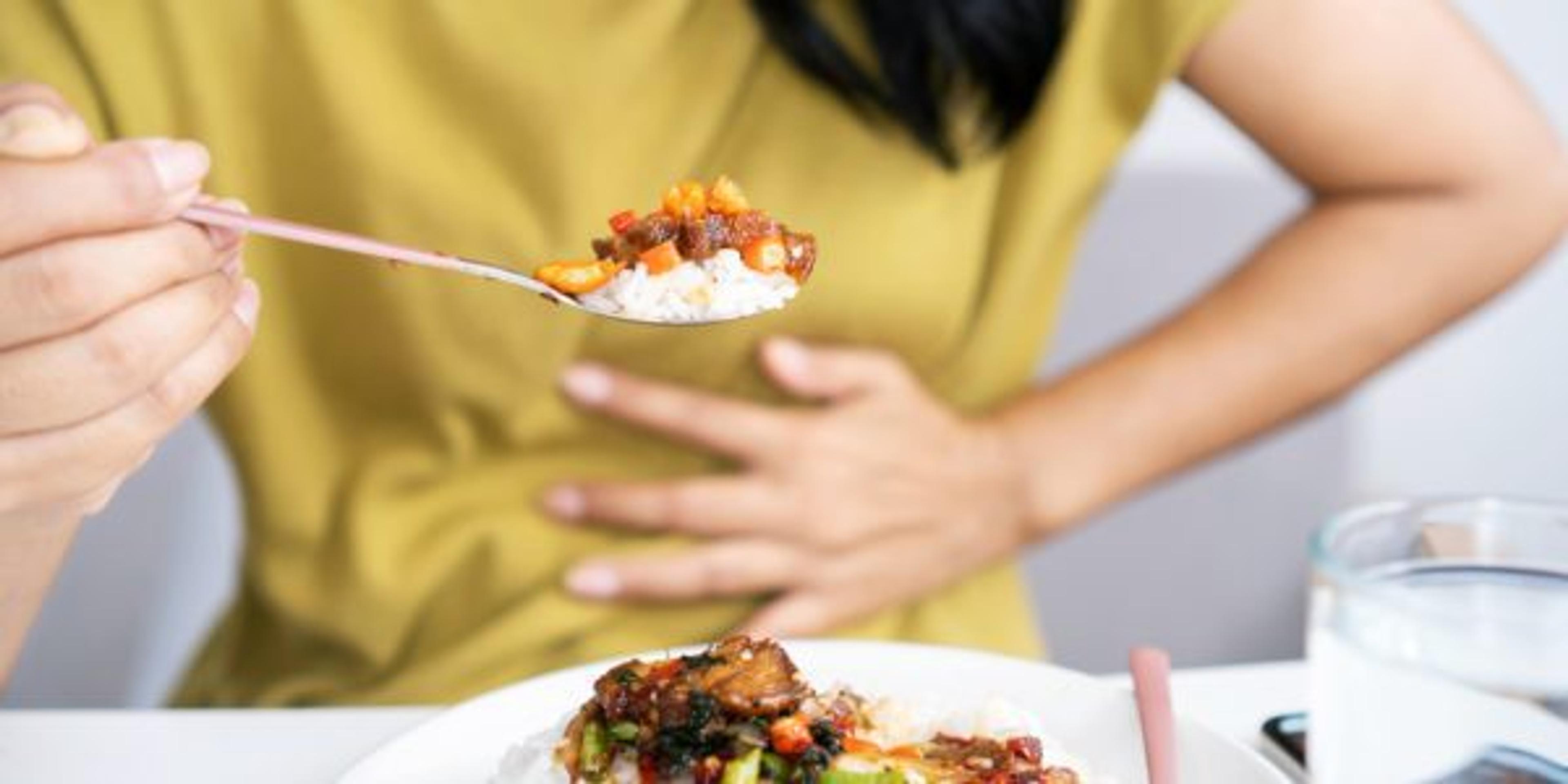 Asian woman eating spicy food and having acid reflux or heartburn hand holding a spoon with chili peppers another hand holding her stomach