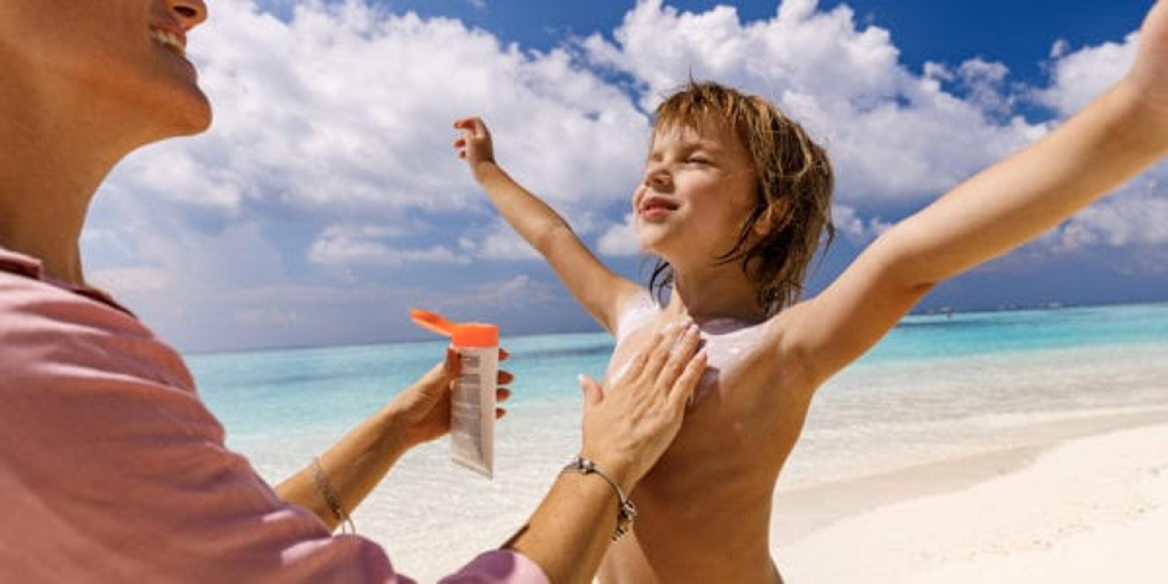 Single mother taking care of her small boy while applying moisturizer on his body during summer day on the beach.