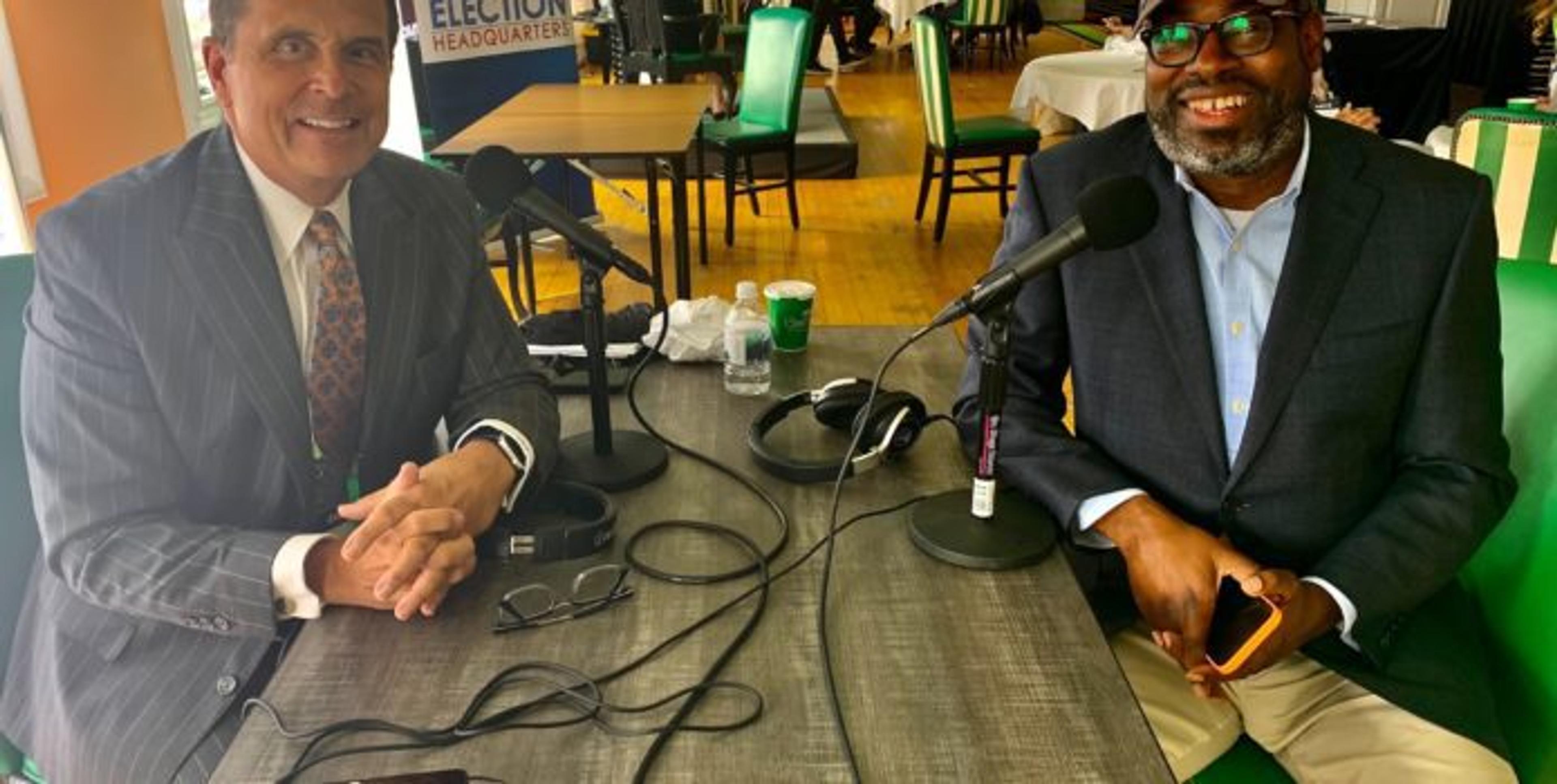 Chuck Gaidica and Stephen Henderson sit at a long, gray table in the main dining room of the Grand Hotel smiling towards the camera.