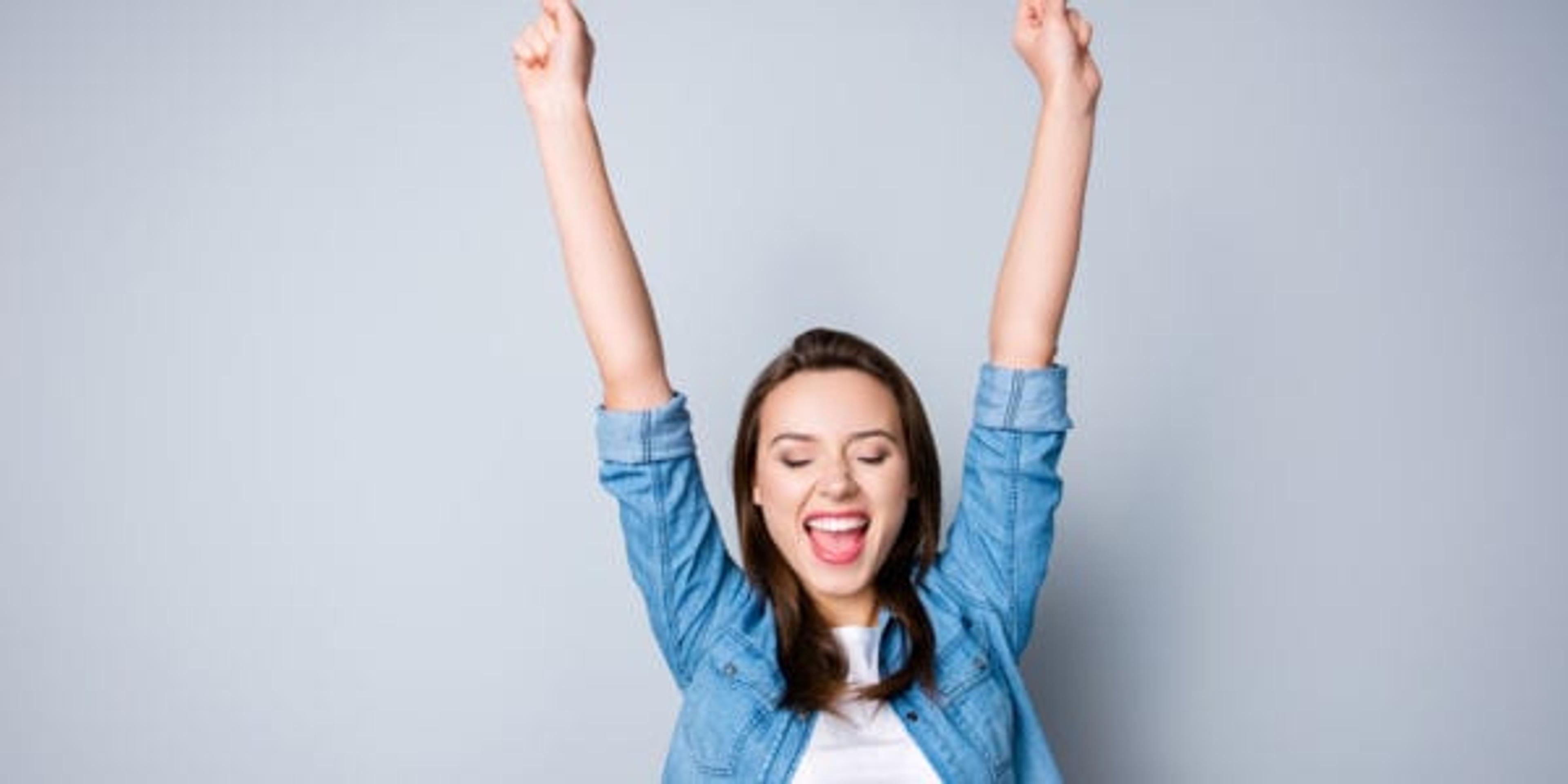 Amazed brunette young business woman in casual shirt is gesturing victory with her raised hands, she is shocked, extremely happy, with closed eyes, beaming smile, open mouth on grey background