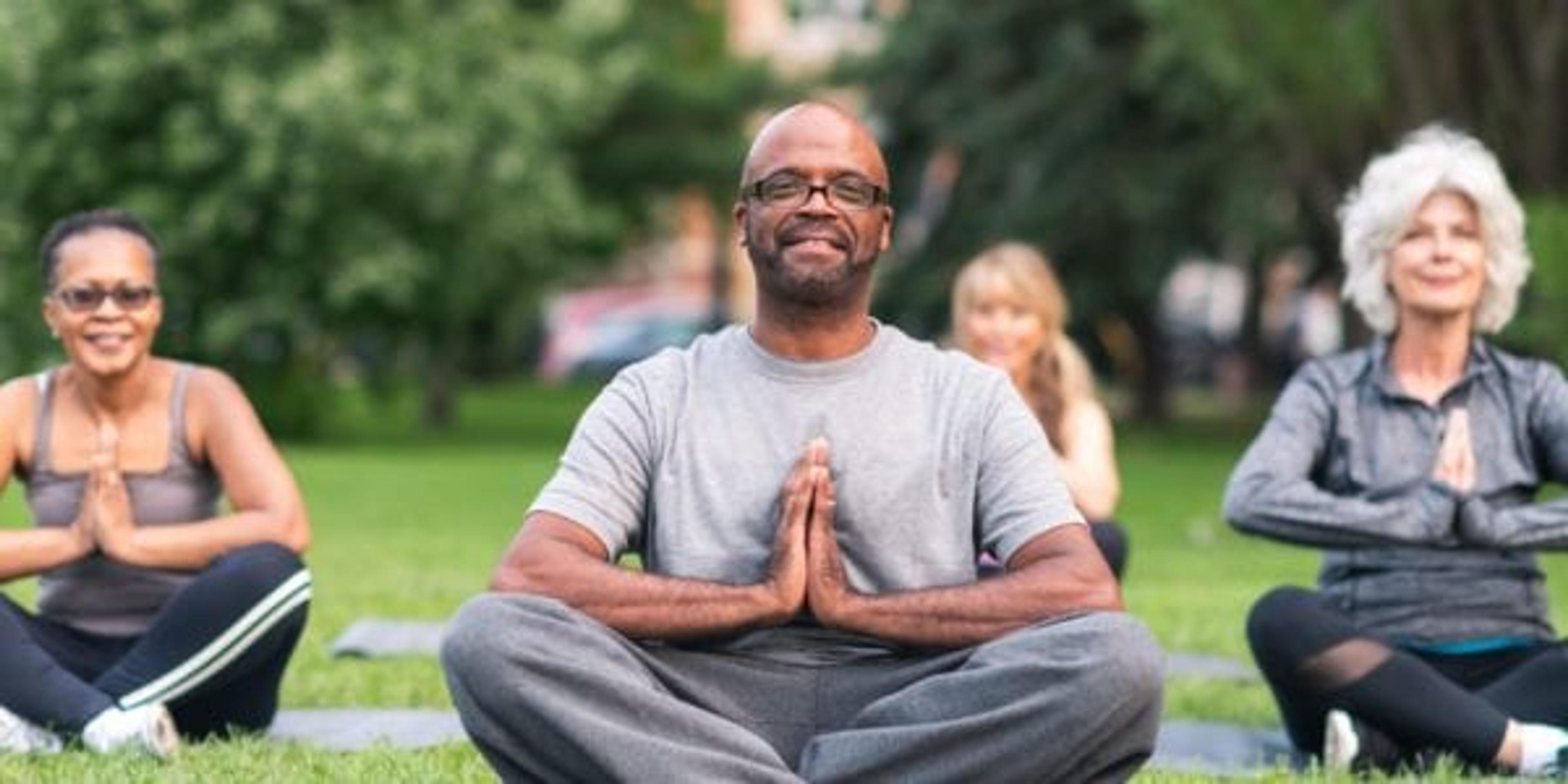 A multi-ethnic group of seniors is attending a yoga class outdoors. The group is sitting on yoga mats. They are meditating. The individual in focus is a black man. He is sitting at the front of the group. He is smiling directly at the camera.
