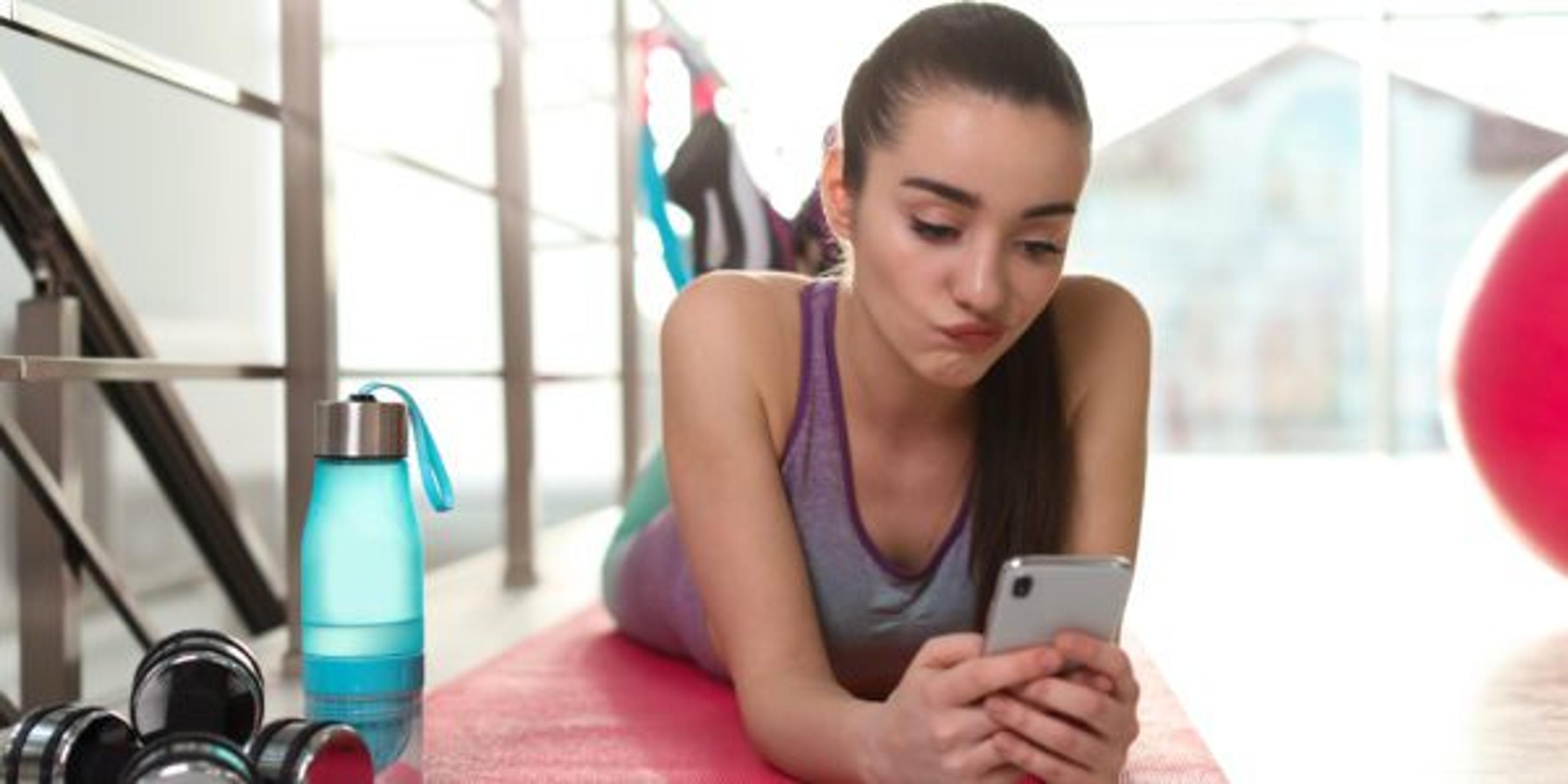 Young woman plays on her smartphone on a yoga mat indoors