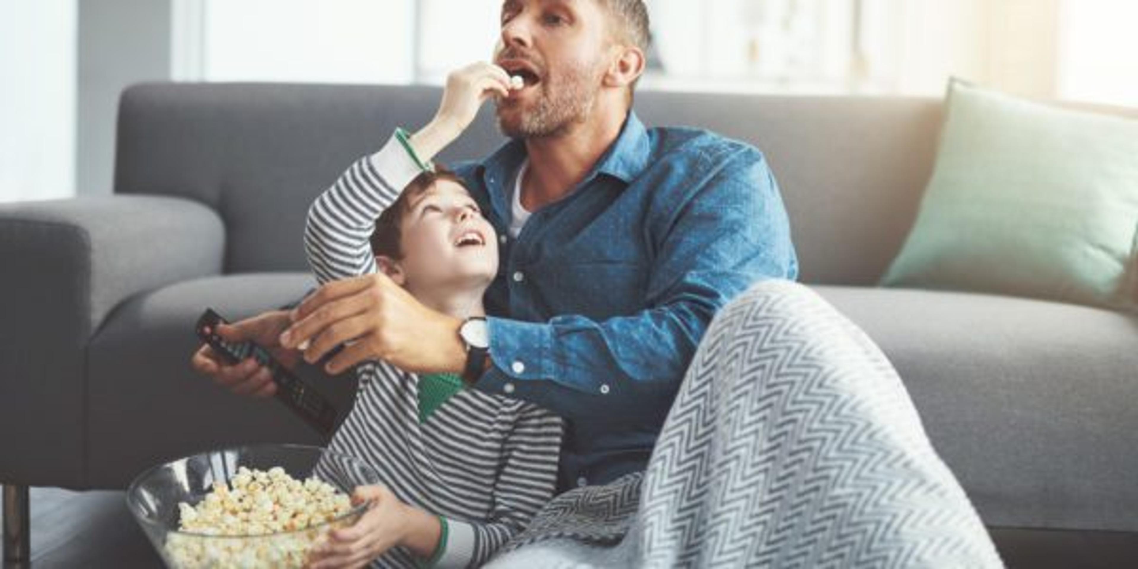 Shot of a carefree young boy and his father watching a movie together while being seated on the floor and eating popcorn at home during the day