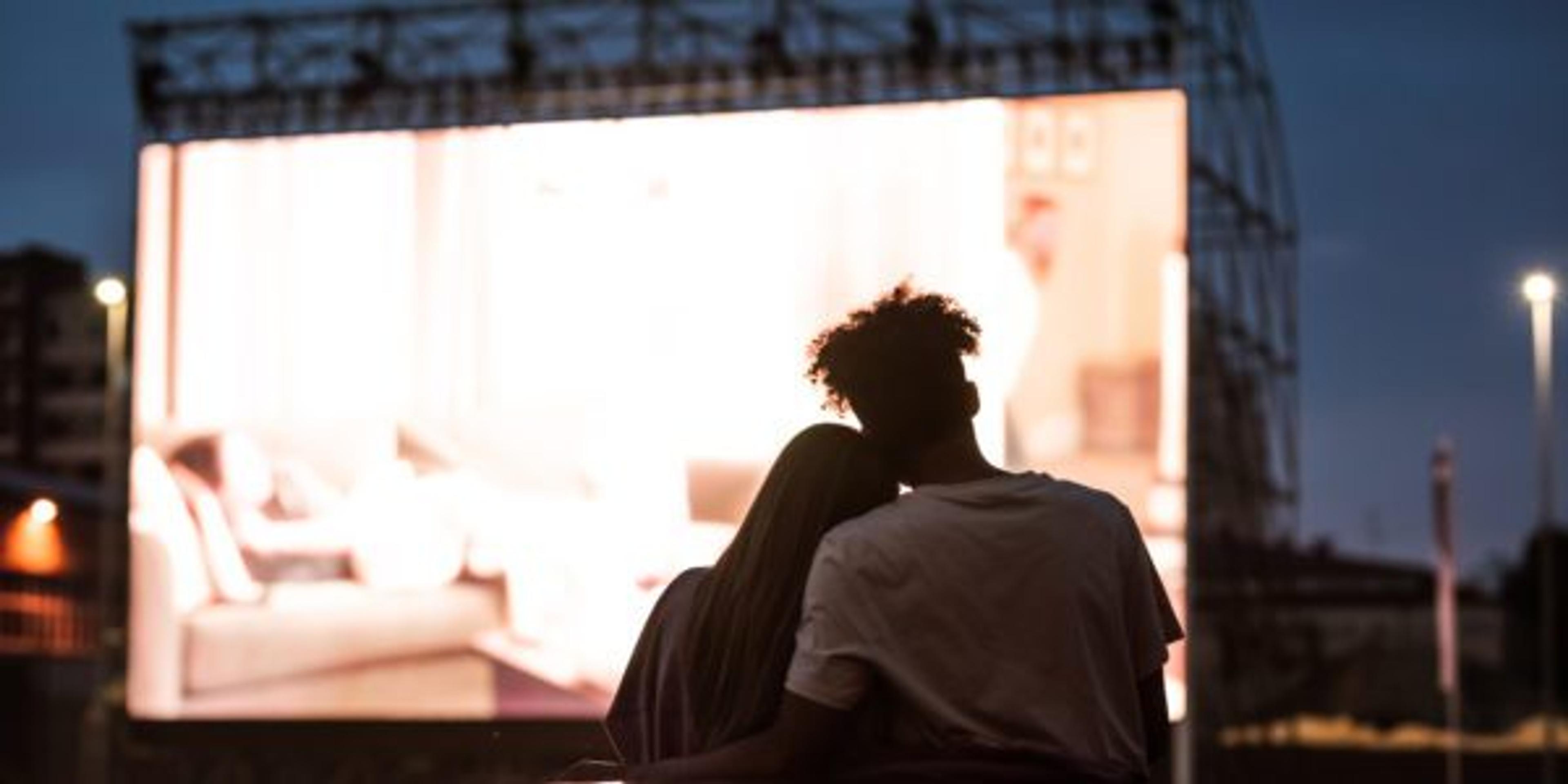 Silhouetted view of attractive young couple, boy and girl embracing, spending time together, sitting in the car while watching a movie in a drive in cinema