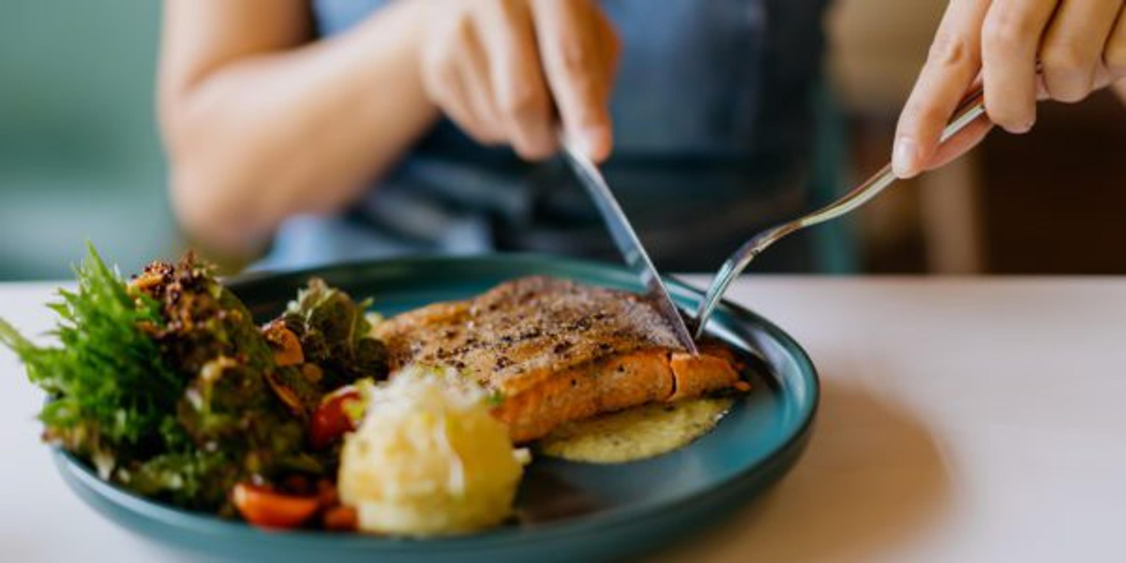 Close up shot of a woman eating pan-fried salmon with a knife and fork in a cafe.
