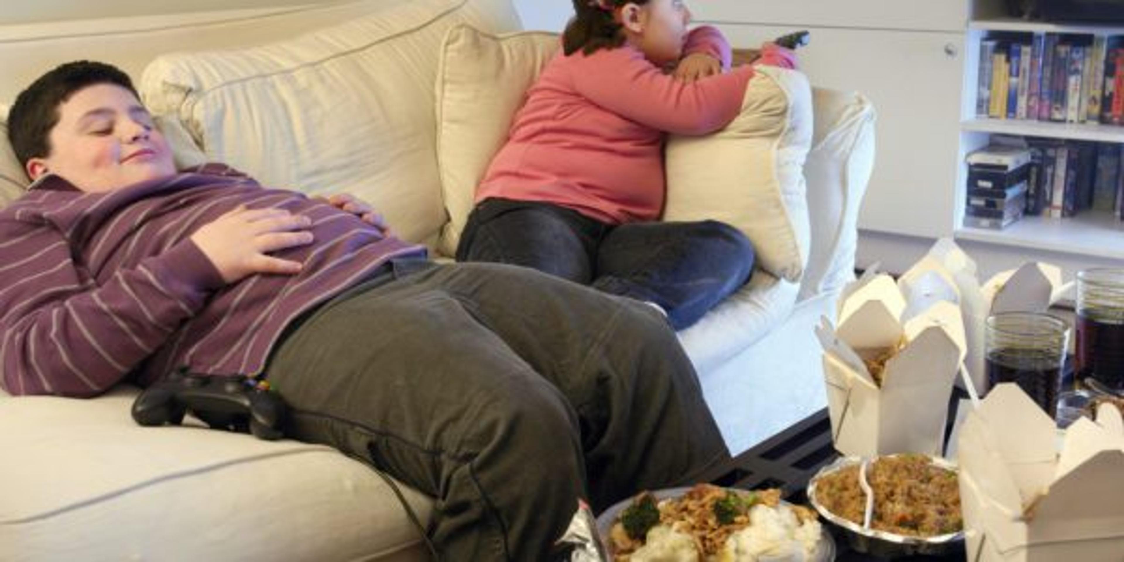 Overweight Brother and Sister Sitting on a Sofa Eating Takeaway Food and Watching the TV