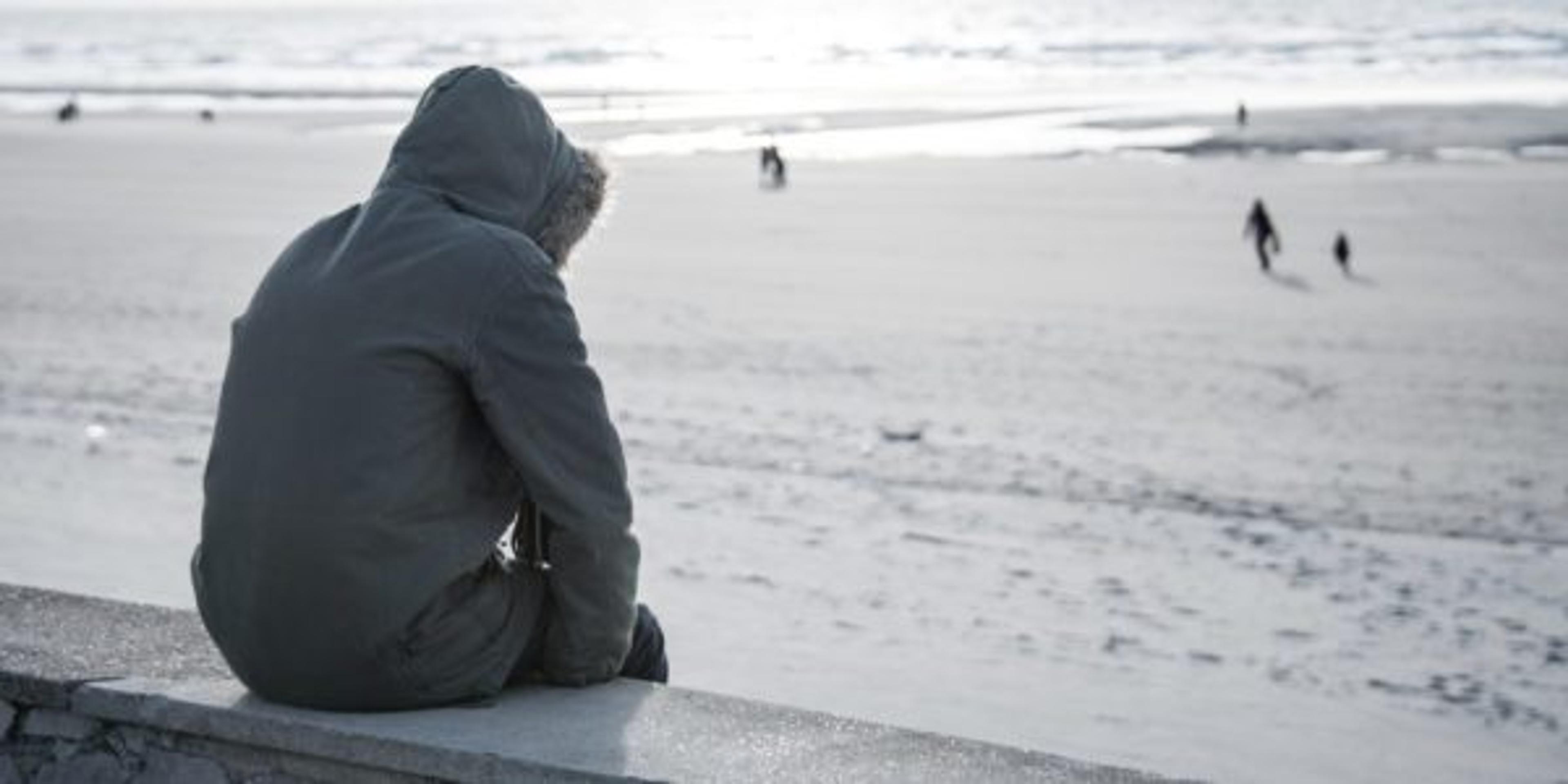 A person wearing a parka coat sits on a wall looking out at the sea