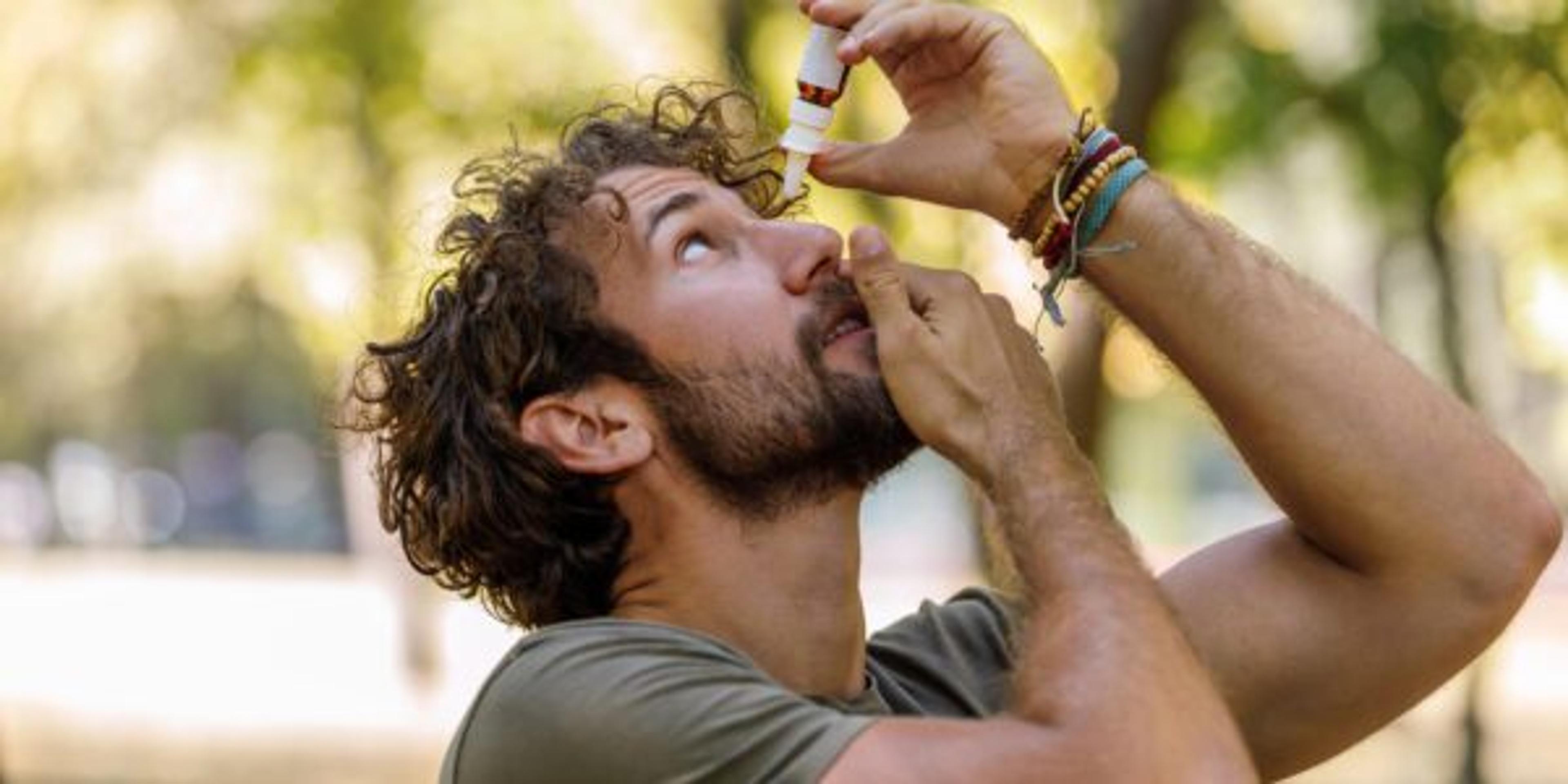 A young adult man going for a walk outdoors stops to apply eyedrops.