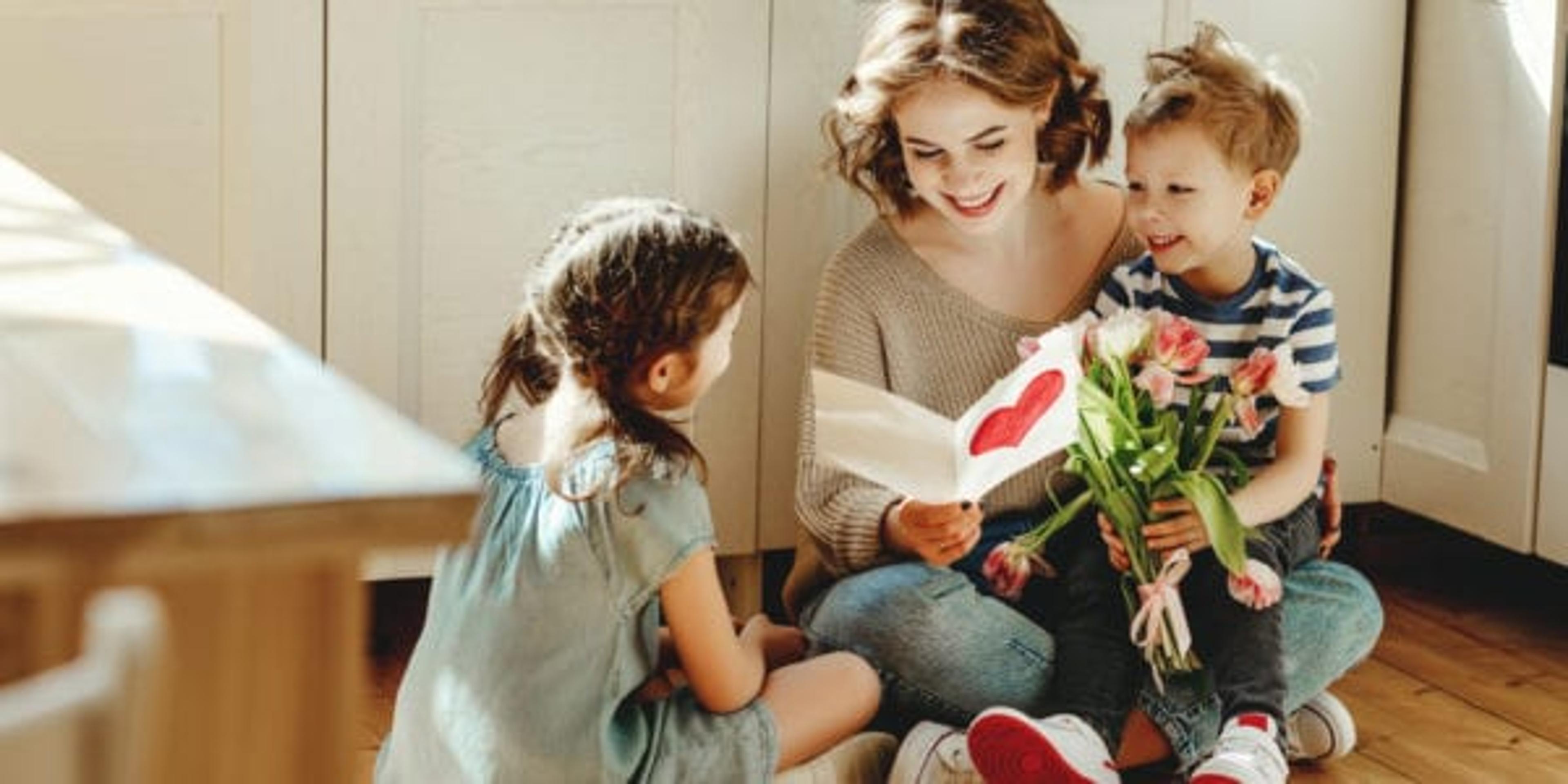 Smiling young woman reading greeting card while sitting on floor with cheerful little siblings with bouquet of flowers during mother day