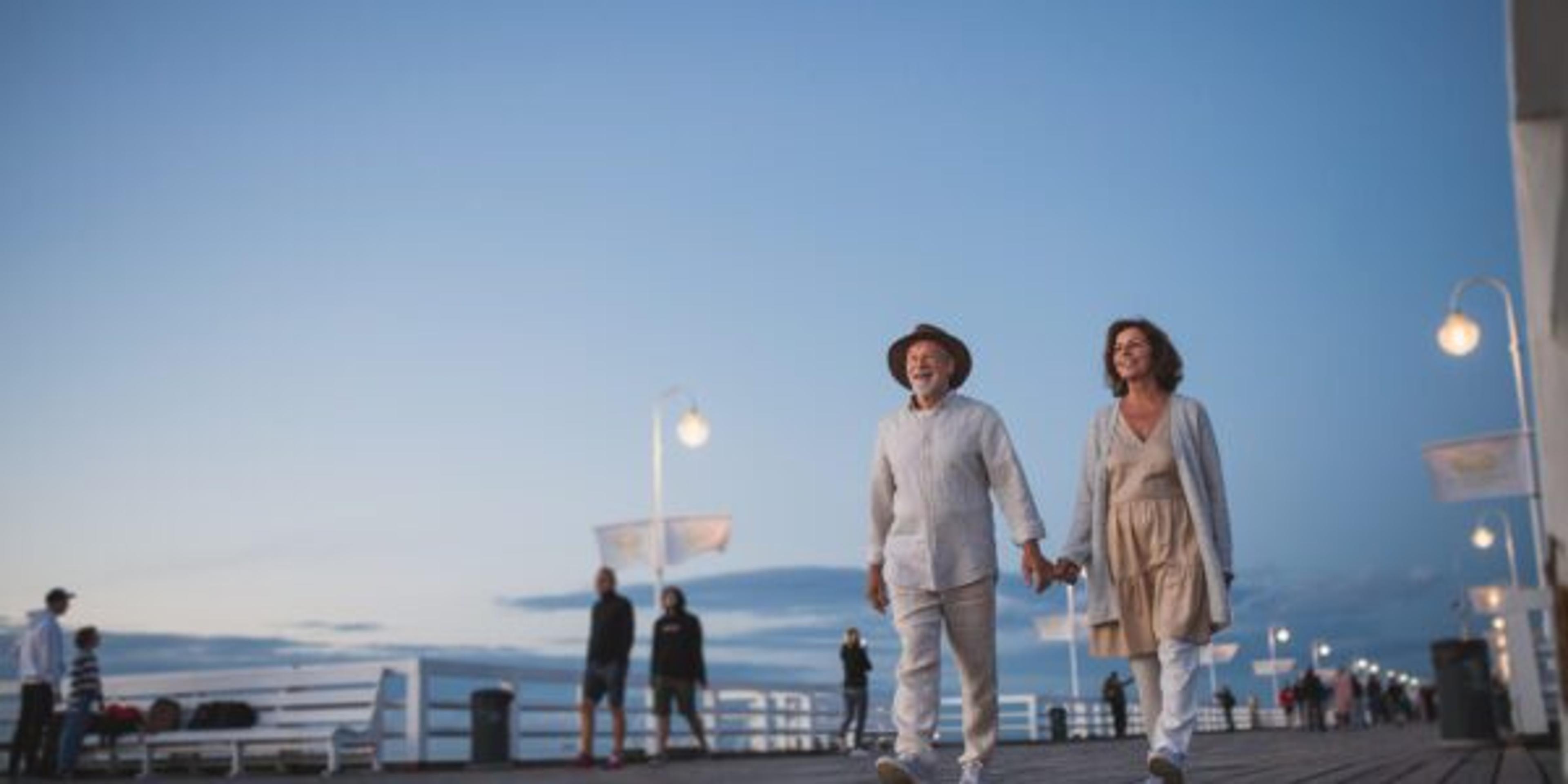 Low angle view of happy senior couple walking outdoors on pier by sea at dusk, holding hands.