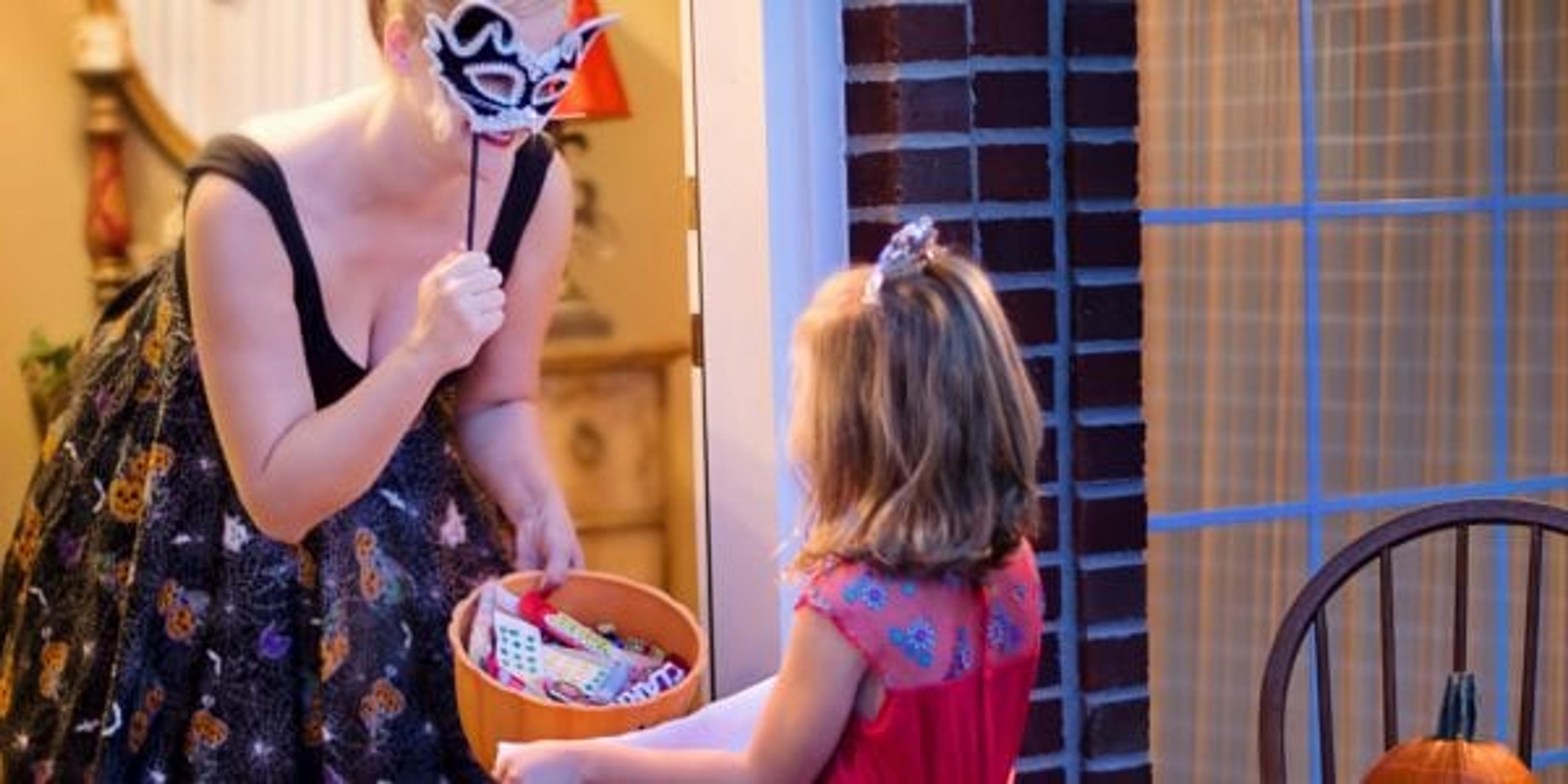 a woman in a black dress holding a mask giving candy to a girl in a pink dress.