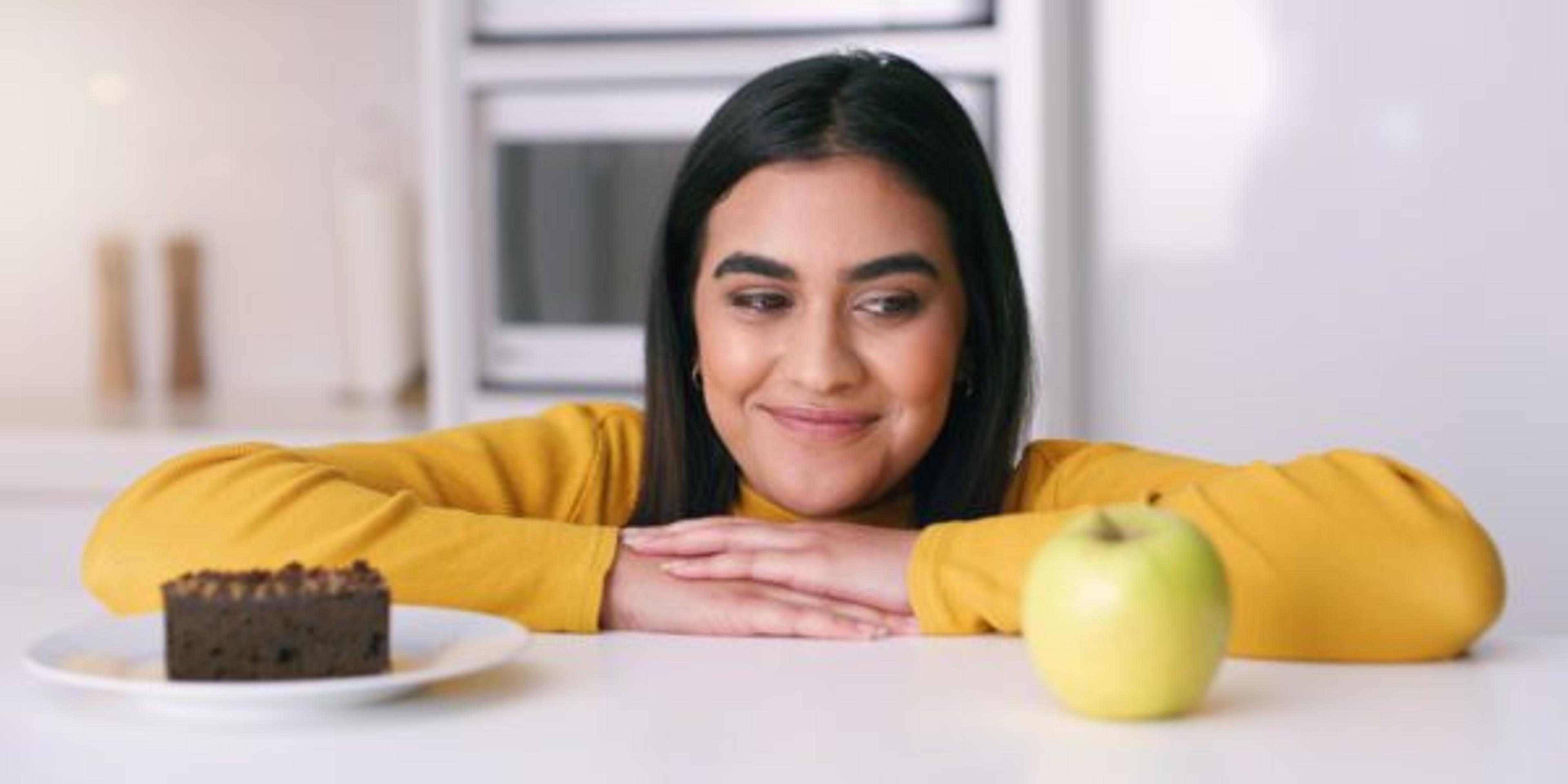 A woman stands at a countertop and stares at both a slice of chocolate cake and an apple.