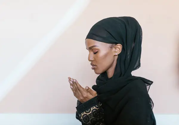 A woman in hijab praying with her hands raised, eyes closed