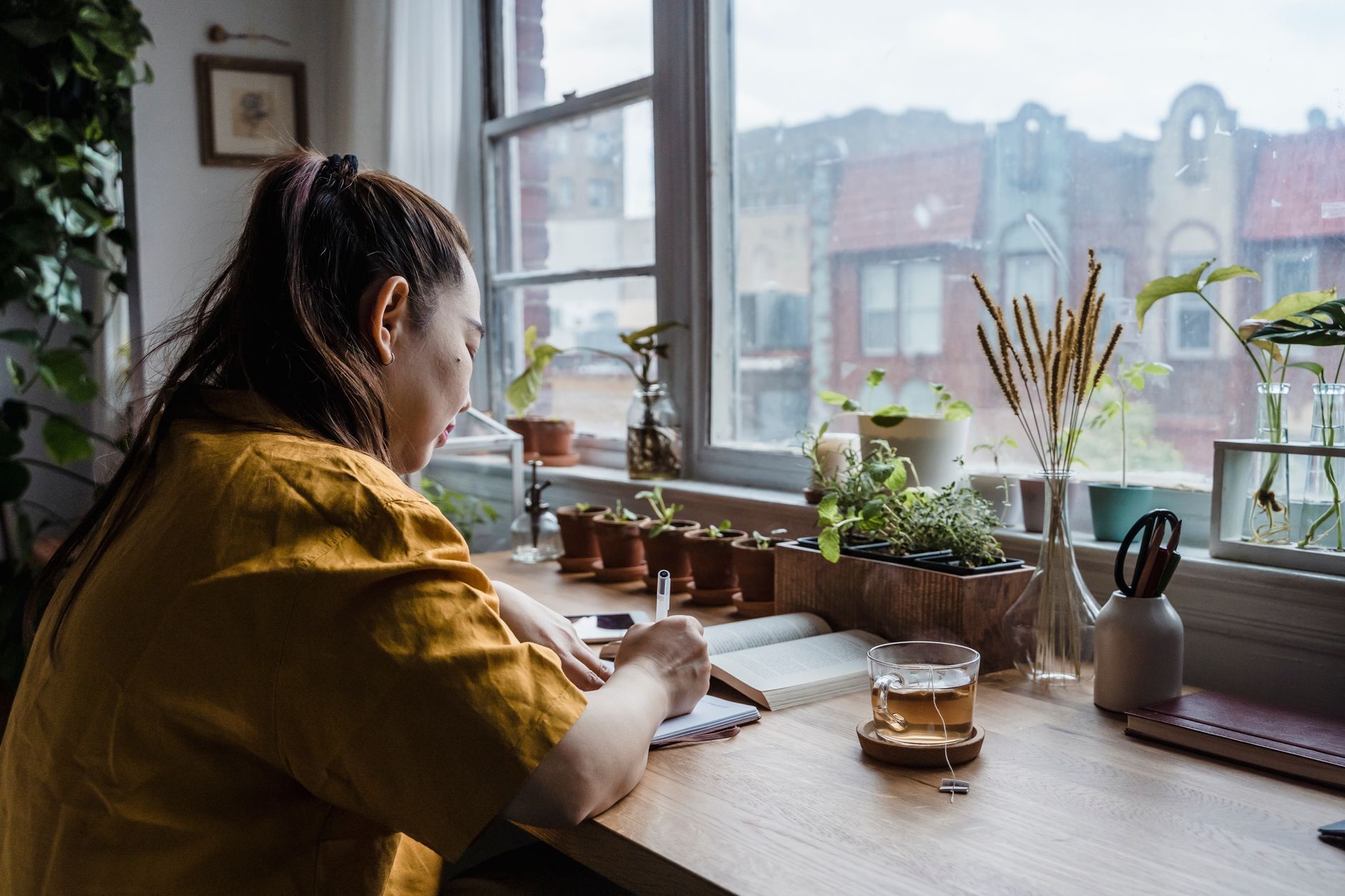 A woman writing in a journal at a sunlit desk full of plants