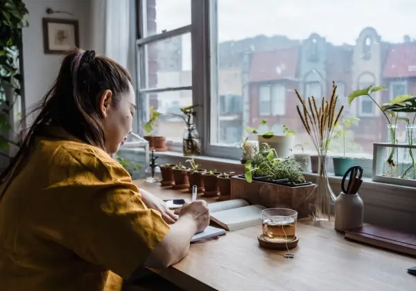 A woman writing in a journal at a sunlit desk full of plants