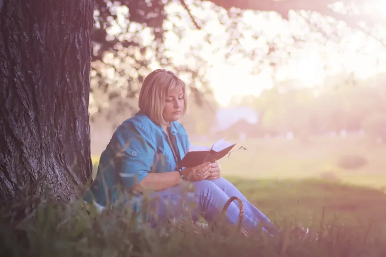 A woman reading quietly at sunset in a park — Scripture as the slow, repeated soil in which the Father heals what was distorted by an earthly father, in his own time.
