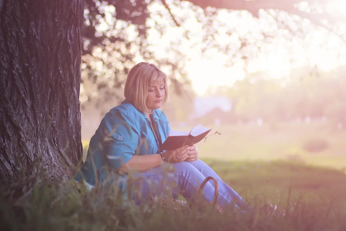 A woman reading quietly at sunset in a park — Scripture as the slow, repeated soil in which the Father heals what was distorted by an earthly father, in his own time.
