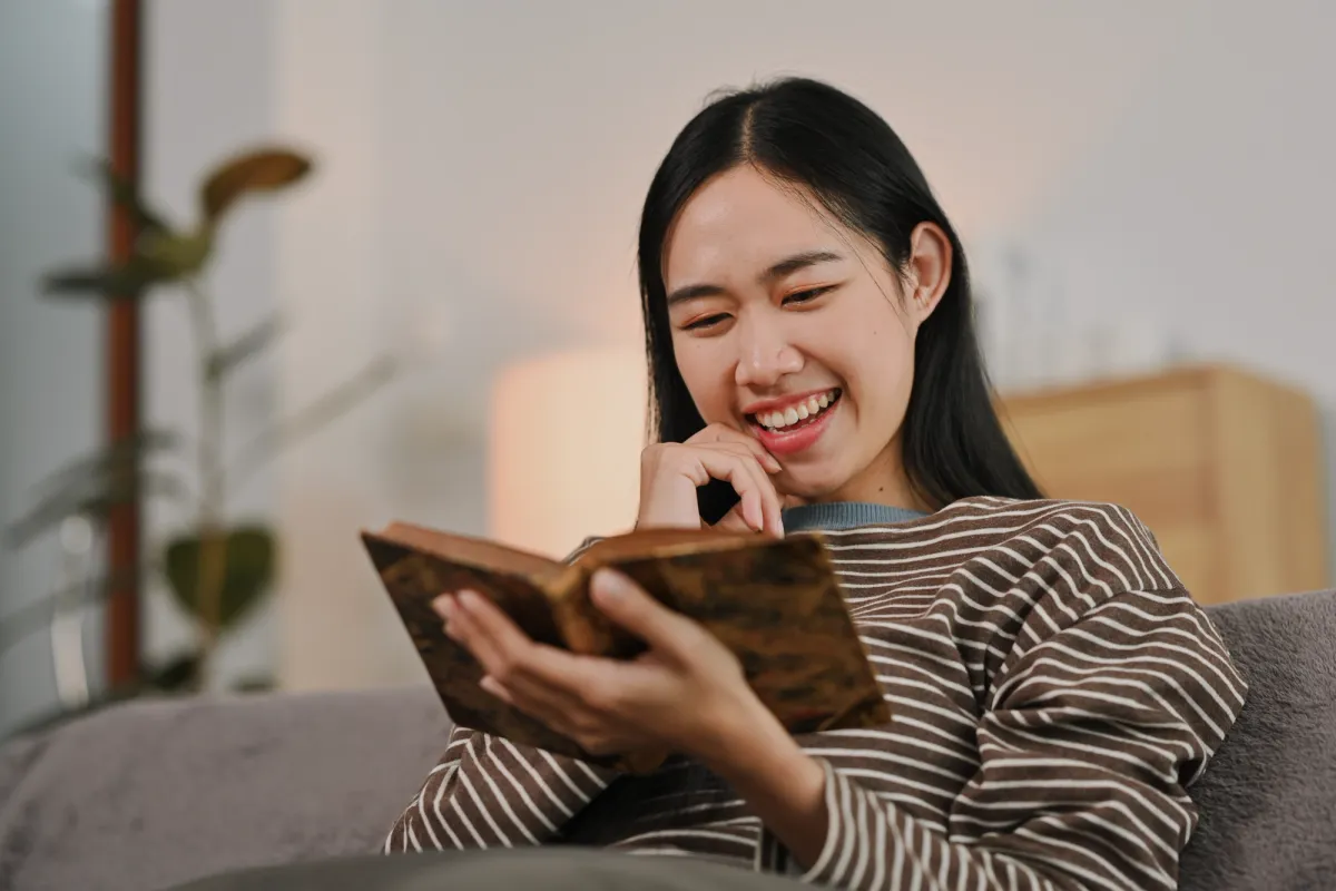 A young woman smiling as she reads Scripture at home — the healing journey from orphan to son begins by letting our Father's Word land in unhurried time.