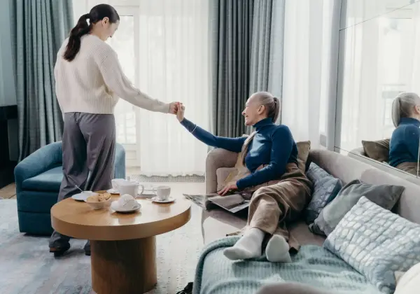 A caregiver gently helping an older woman up from the sofa