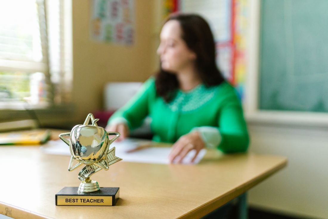 A 'Best Teacher' trophy on a desk beside a weary teacher