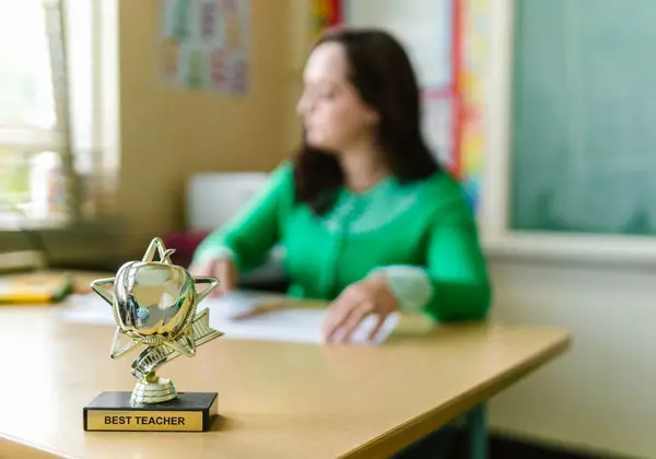 A 'Best Teacher' trophy on a desk beside a weary teacher