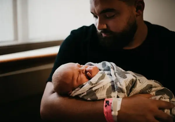 A father gently holding his sleeping newborn by a window