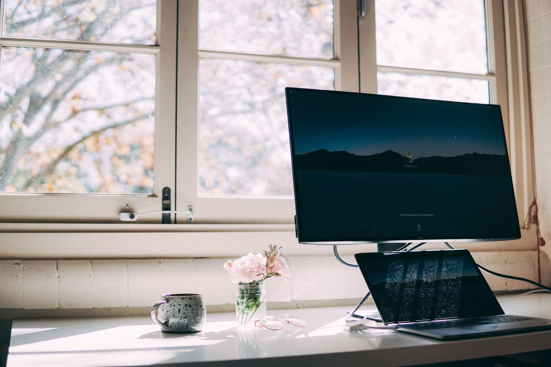 A tidy desk by a bright window with a coffee mug and flowers