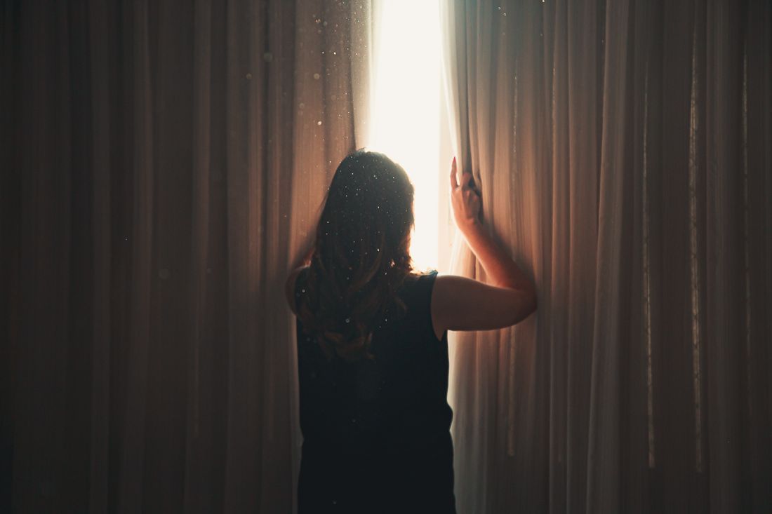 A woman drawing back the curtain to let in the morning light