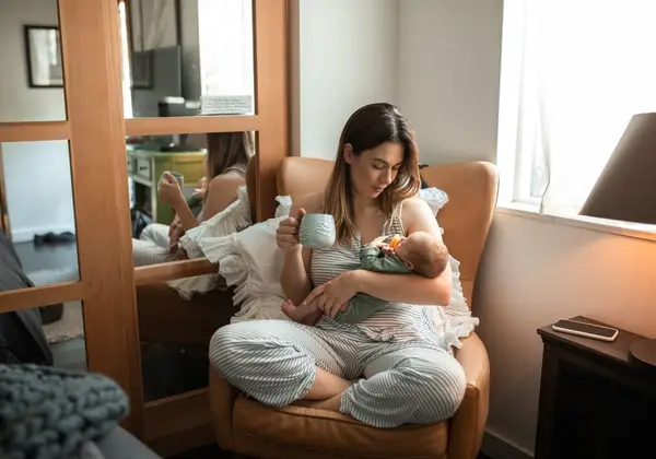A mother nursing her baby with a cup of coffee in a quiet chair